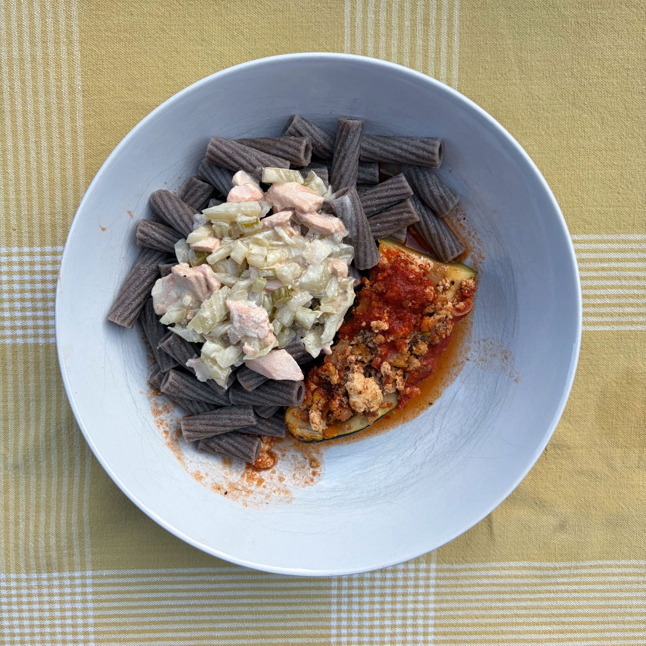 Weißer tiefer Teller mit Buchweizen-Rigatoni, belegt mit zwei Portionen Reste: links eine helle Lachssoße mit Zwiebeln, rechts ein Tomaten-Hackfleisch-Sugo auf einer Zucchinischeibe. Der Teller steht auf einem gelb-weiß karierten Tischtuch.