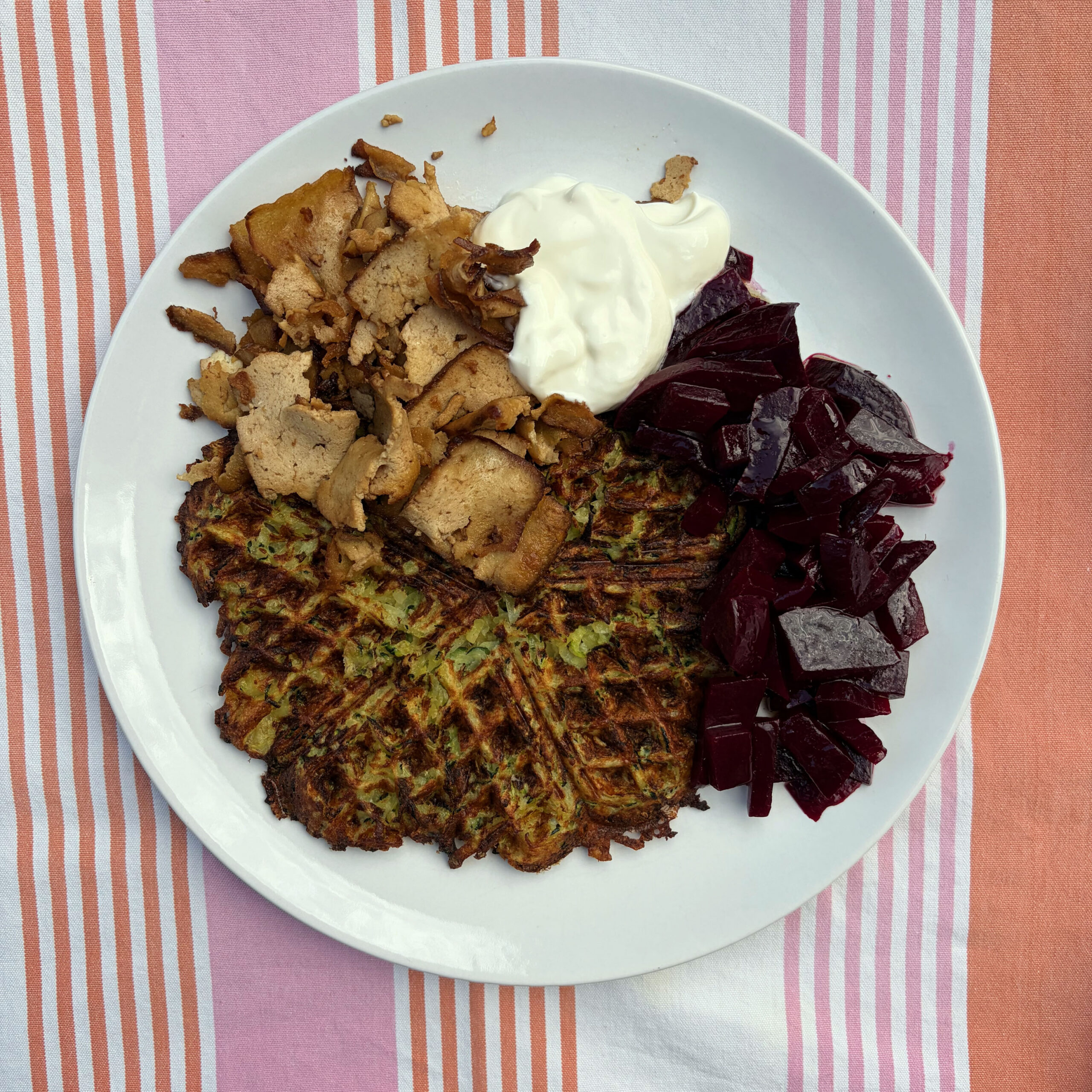 Zwei herzhafte Zucchini-Kartoffel-Waffeln auf einem weißen Teller, belegt mit gebratenen Räuchertofu-Scheiben und einem Klecks weißem Joghurt, daneben ein Rote-Bete-Salat mit Balsamico. Der Teller liegt auf einem rosa-orangefarbenen Streifentuch.