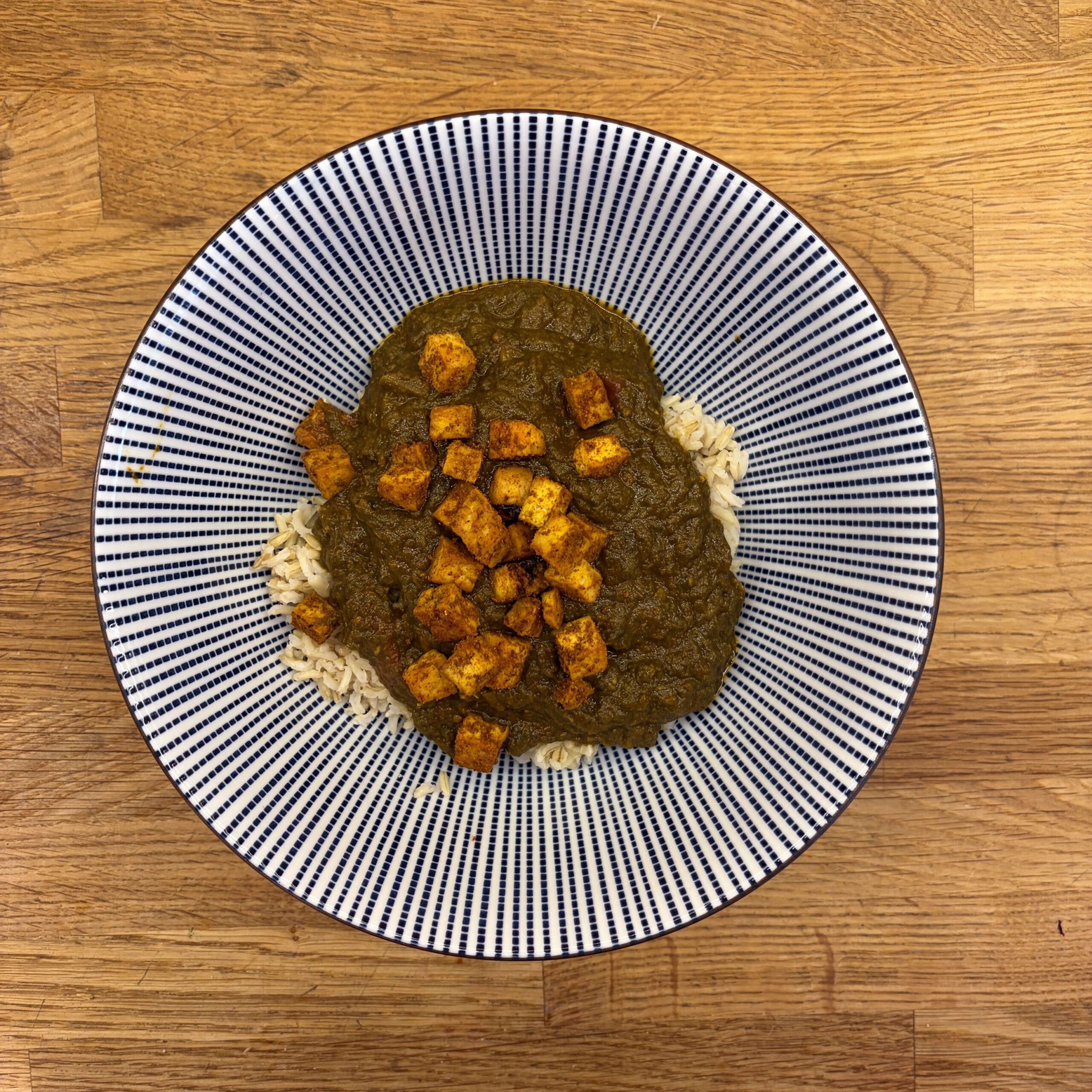 Palak Tofu mit Vollkornreis: Gebratene, gewürzte Tofuwürfel auf dunkelgrüner Spinat-Tomaten-Sauce, serviert auf Vollkornreis, angerichtet auf einem weiß-blau gestreiften Teller.
