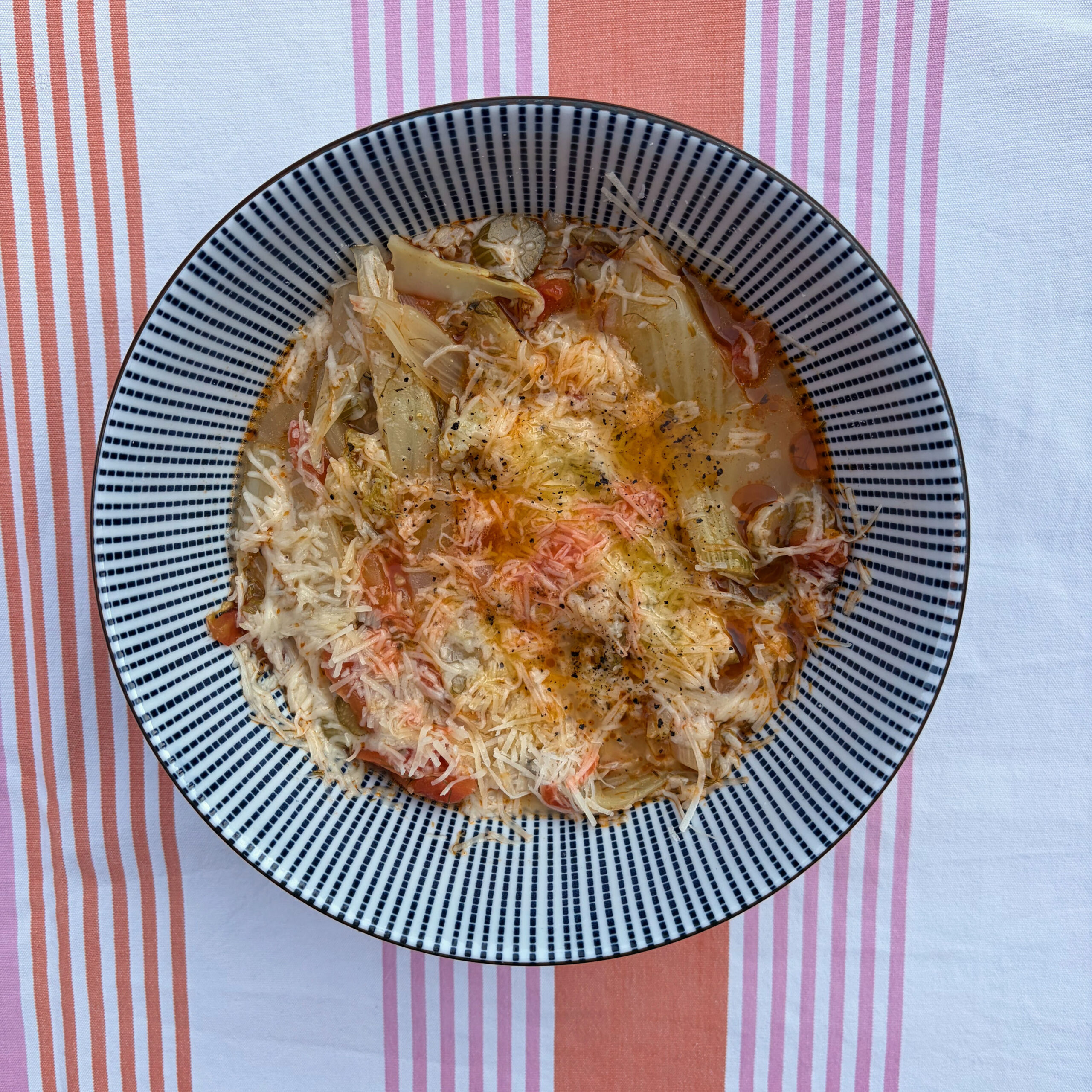 Tiefer Teller mit Fenchel-Ragout, bestehend aus geschmortem Fenchel in Tomatensoße, großzügig mit geriebenem Parmesan bestreut und mit schwarzem Pfeffer gewürzt. Der Teller hat ein schwarz-weißes Streifenmuster und steht auf einer rosa-weiß gestreiften Tischdecke.