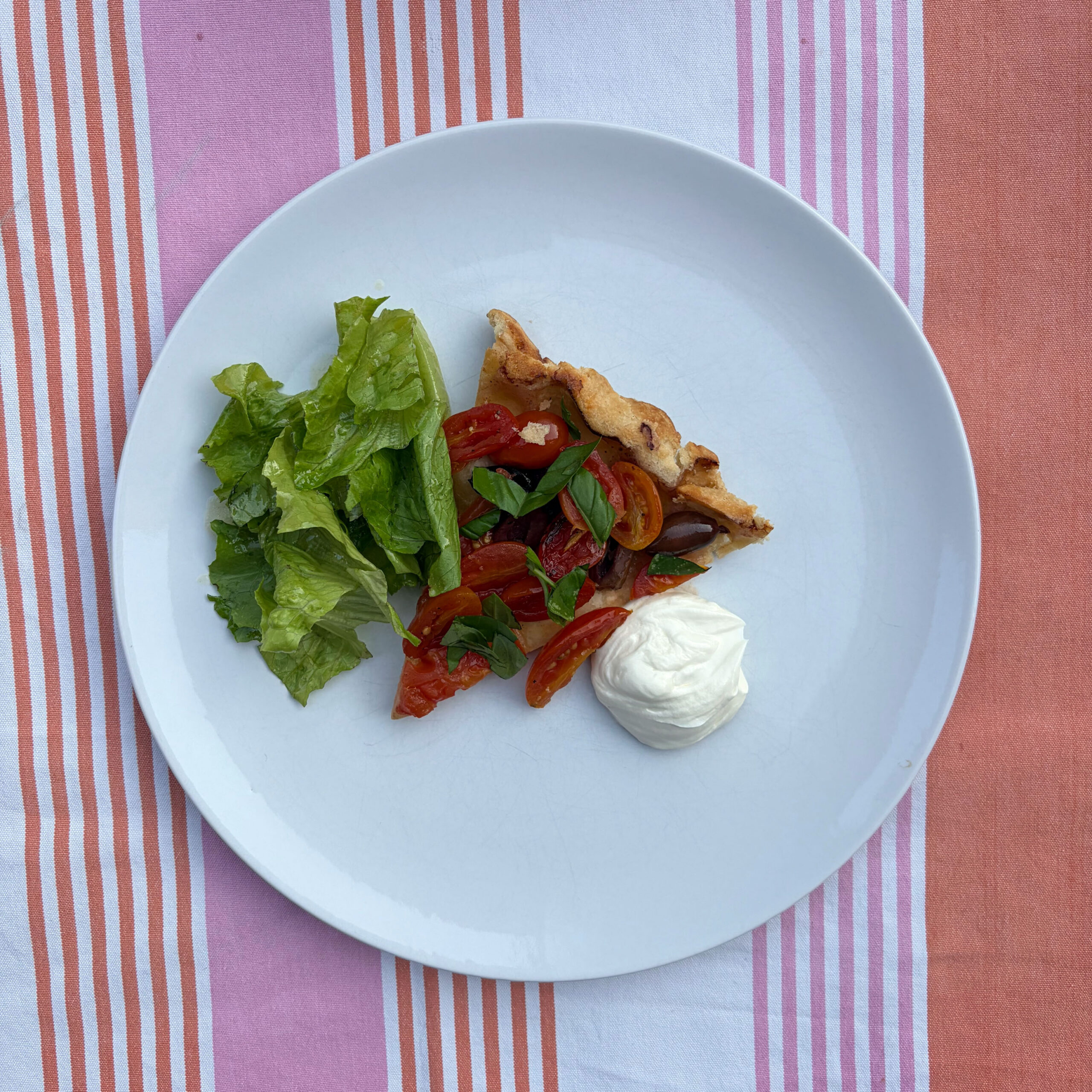 Ein Stück Tomaten-Tarte-Tatin mit karamellisierten Kirschtomaten, Kalamata-Oliven und frischem Basilikum auf einem weißen Teller, serviert mit einem Klecks Ziegenfrischkäse-Dip und grünem Blattsalat. Der Teller liegt auf einem rot-weiß-rosa gestreiften Tischtuch.