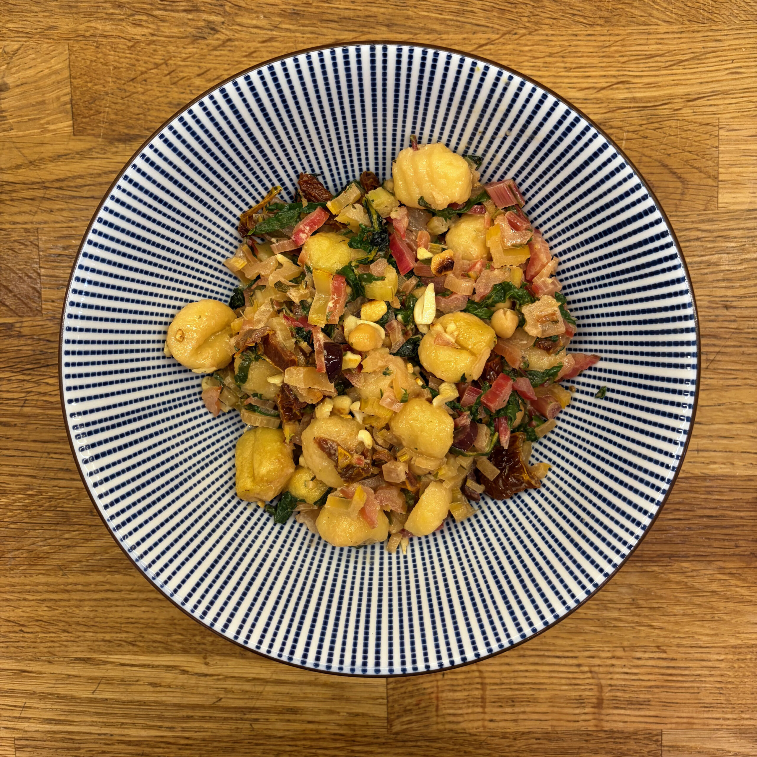 Ein Teller mit Gnocchi-Auflauf mit Mangold, Kichererbsen, Oliven, getrockneten Tomaten und gehackten Cashewkernen, serviert auf einem blau-weiß gestreiften Teller auf einem Holztisch.