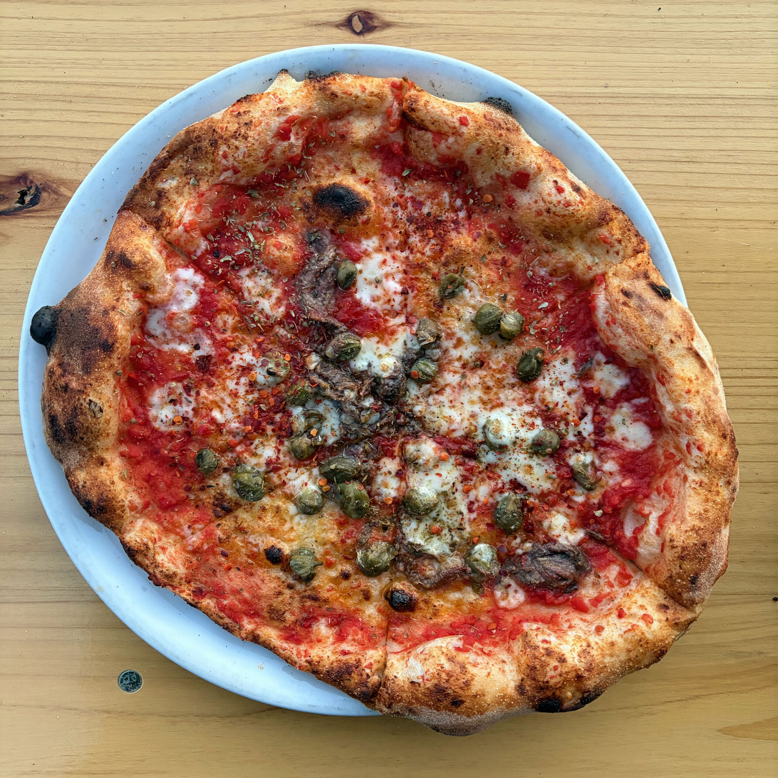 Neapolitanische Pizza Romana auf weißem Teller, belegt mit Tomatensauce, Mozzarella, Sardellen, Kapern und Chiliflakes, mit charakteristisch blasigem, stellenweise verkohltem Rand.