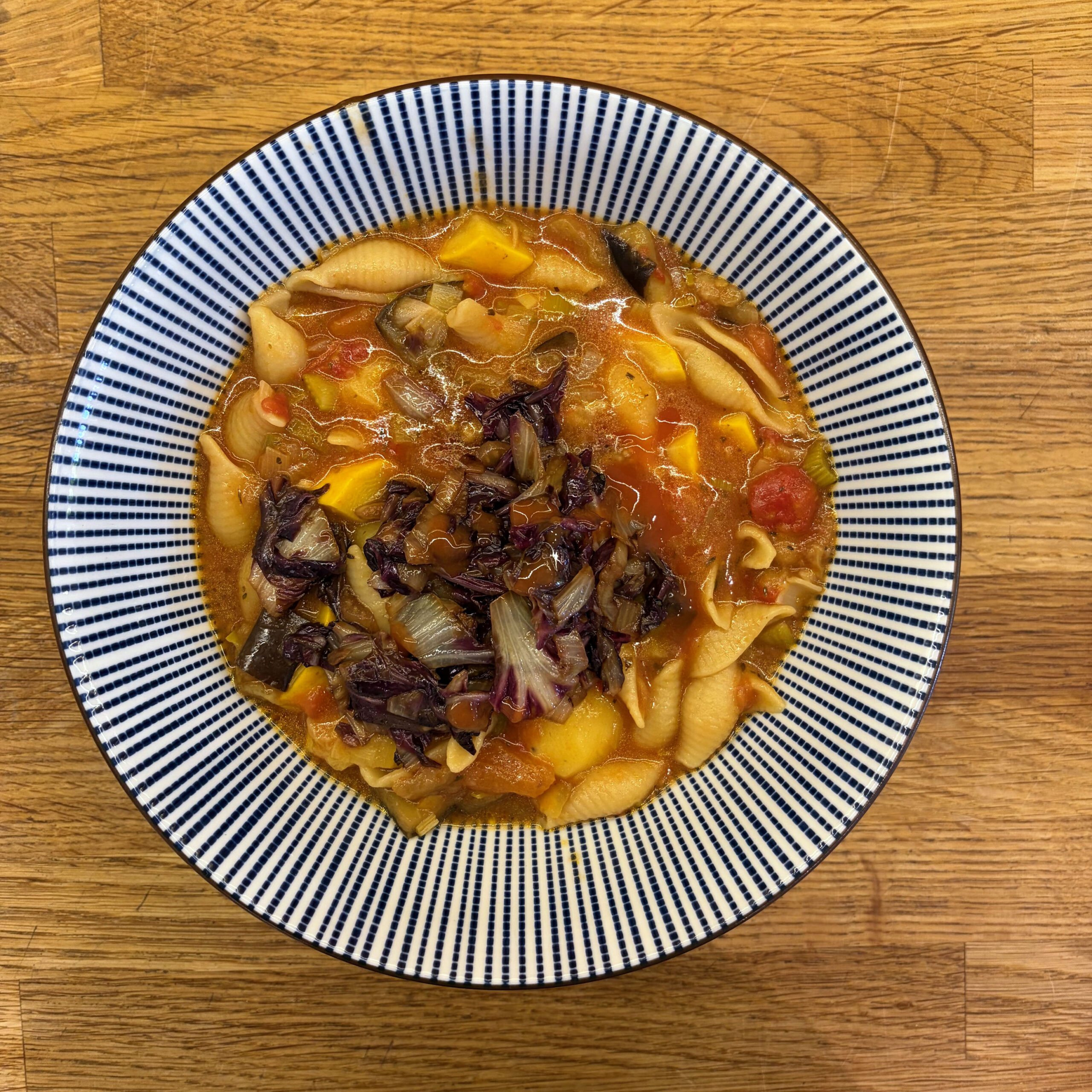 Minestrone-Suppe mit Conchiglie-Nudeln, Aubergine, Zucchini, Kartoffeln, Sellerie und Tomaten in einem blau-weiß gestreiften Suppenteller auf Holzuntergrund.