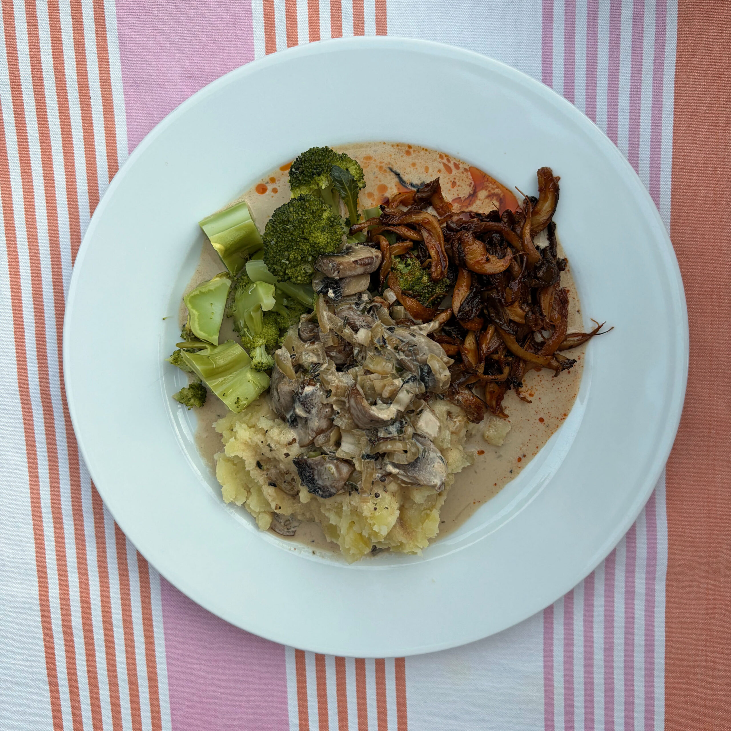 Weißer Teller mit Kartoffelpüree, Champignonsoße, gedämpftem Brokkoli und gebratenen Austernpilzen auf einem rot-weiß gestreiften Tischtuch.