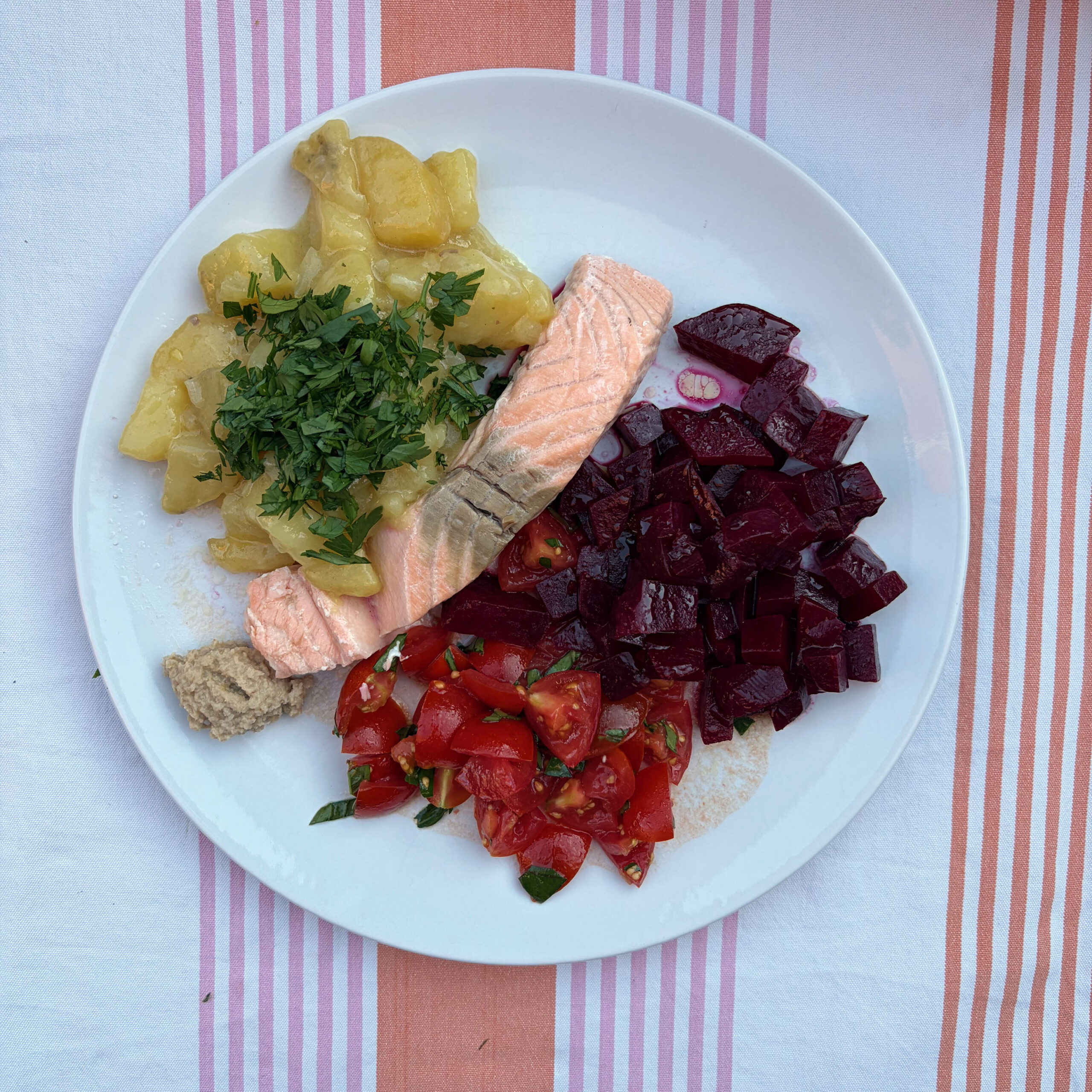Teller mit gedämpftem Lachsfilet, Kartoffelsalat mit gehackter Petersilie, gewürfeltem Rote-Bete-Salat und Tomatenstücken mit Basilikum auf gestreiftem Tischtuch.