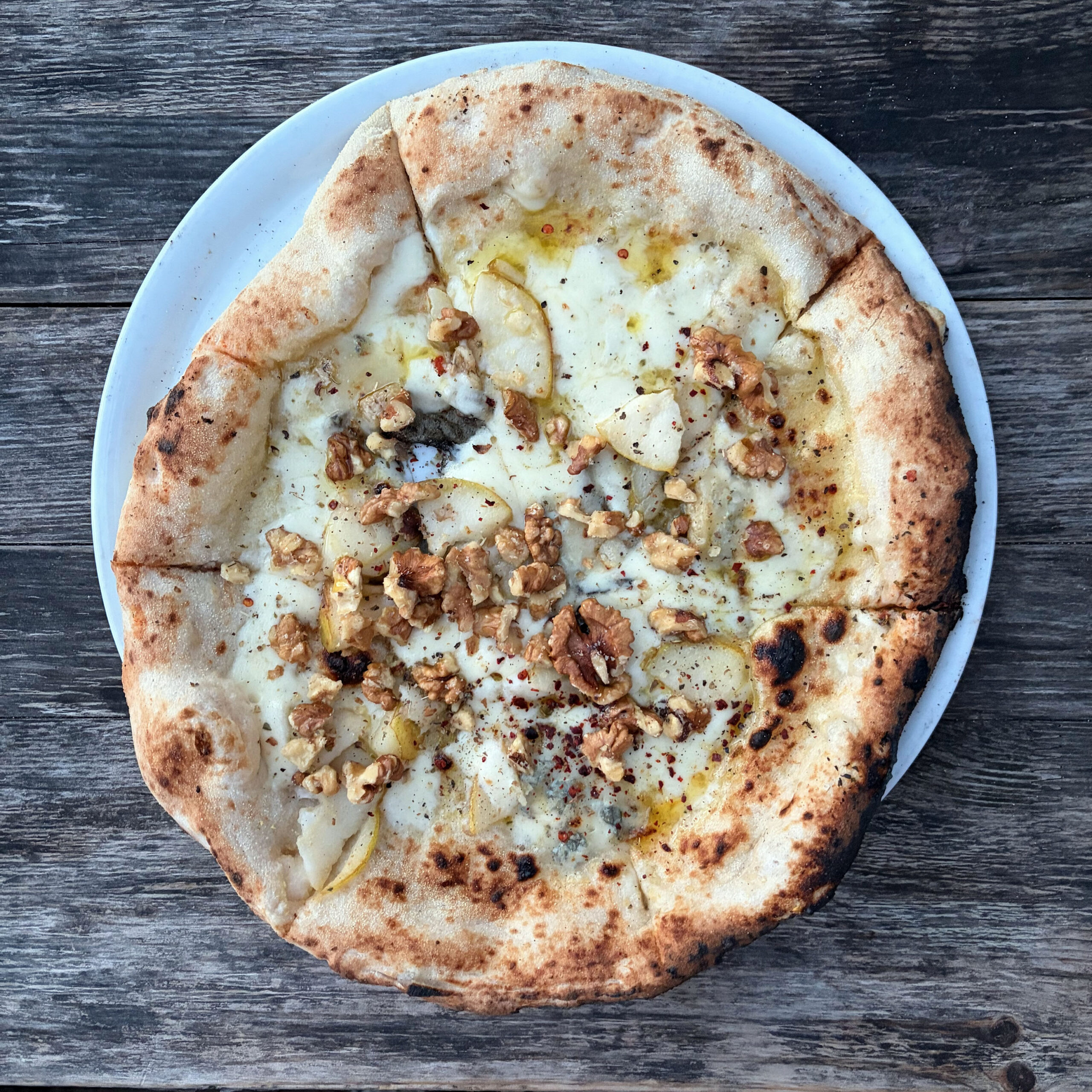 Pizza bianca mit geschmolzenem Käse, Gorgonzola, Walnussstücken und Birnenscheiben auf einem weißen Teller, serviert auf einem Holztisch. Der Teig ist neapolitanisch mit leicht verkohltem Rand.
