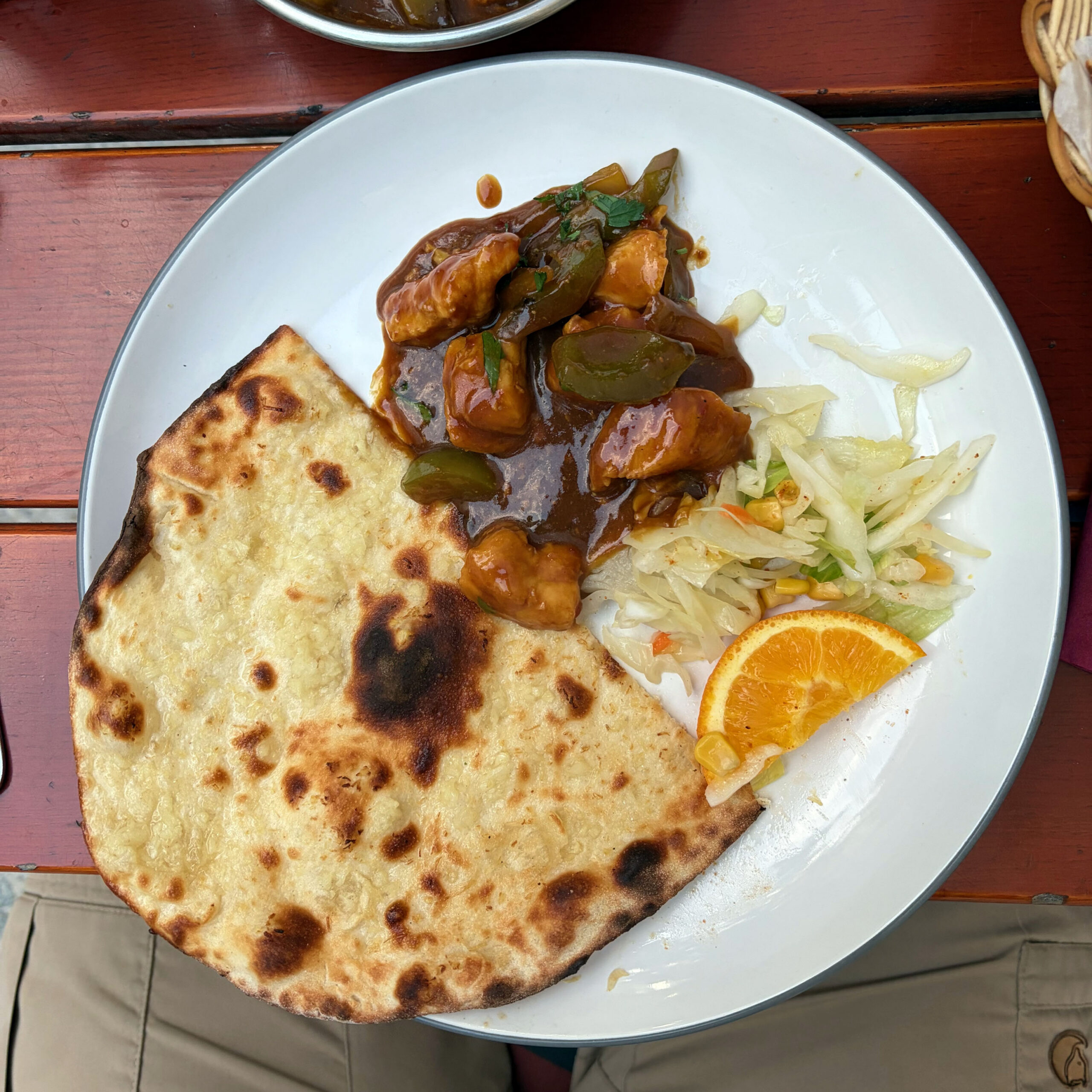 Ein weißer Teller mit Chili Chicken in dunkler Soße mit grünen Paprikastücken, einem großen, leicht verkohlten Knoblauch-Naan sowie einem frischen Krautsalat und einer Orangenscheibe als Beilage.