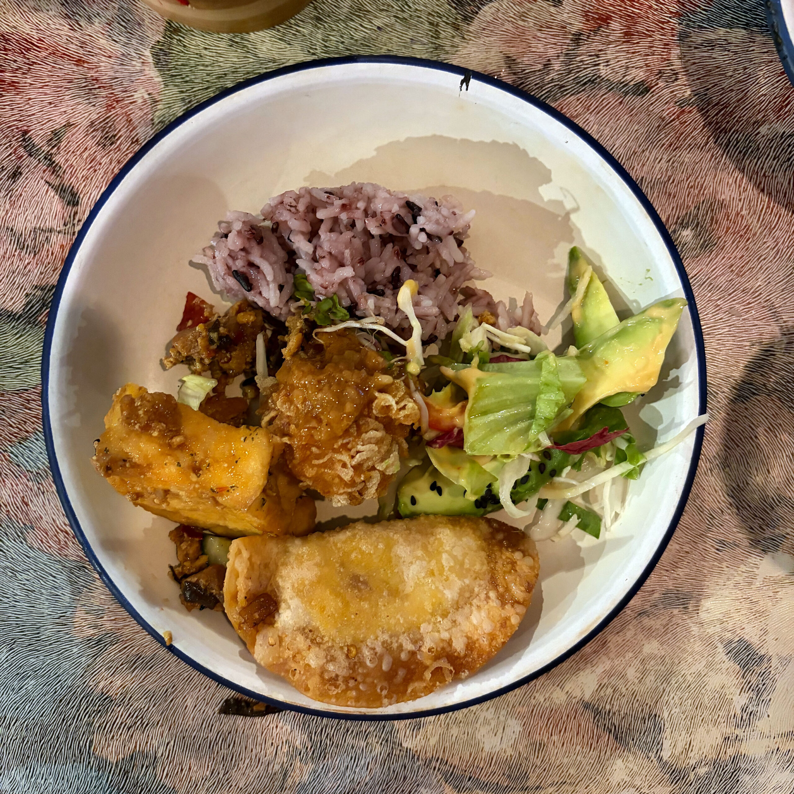 Vegane Thai-Bowl mit lila Reis, frittiertem Tofu, Gemüsecurry, grünem Salat mit Avocado und einem gebratenen Teigtascherl, serviert in einer weißen Schüssel mit blauem Rand.