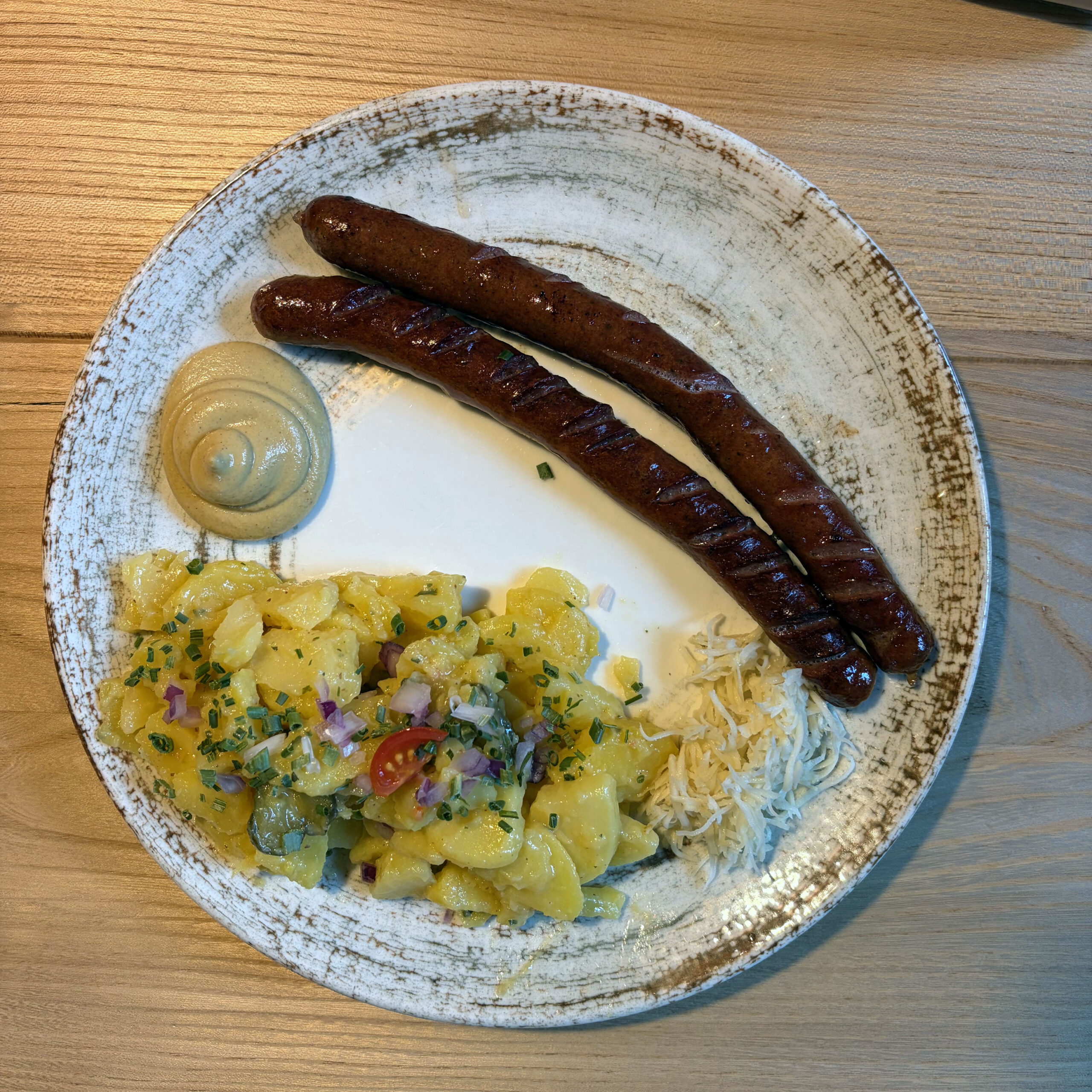 Zwei gegrillte Rindsbratwürste auf einem rustikalen weißen Teller, serviert mit Kartoffelsalat (mit roten Zwiebeln und Schnittlauch), Sauerkraut und einem kleinen Dip, auf einem Holztisch im Spitzsteinhaus.