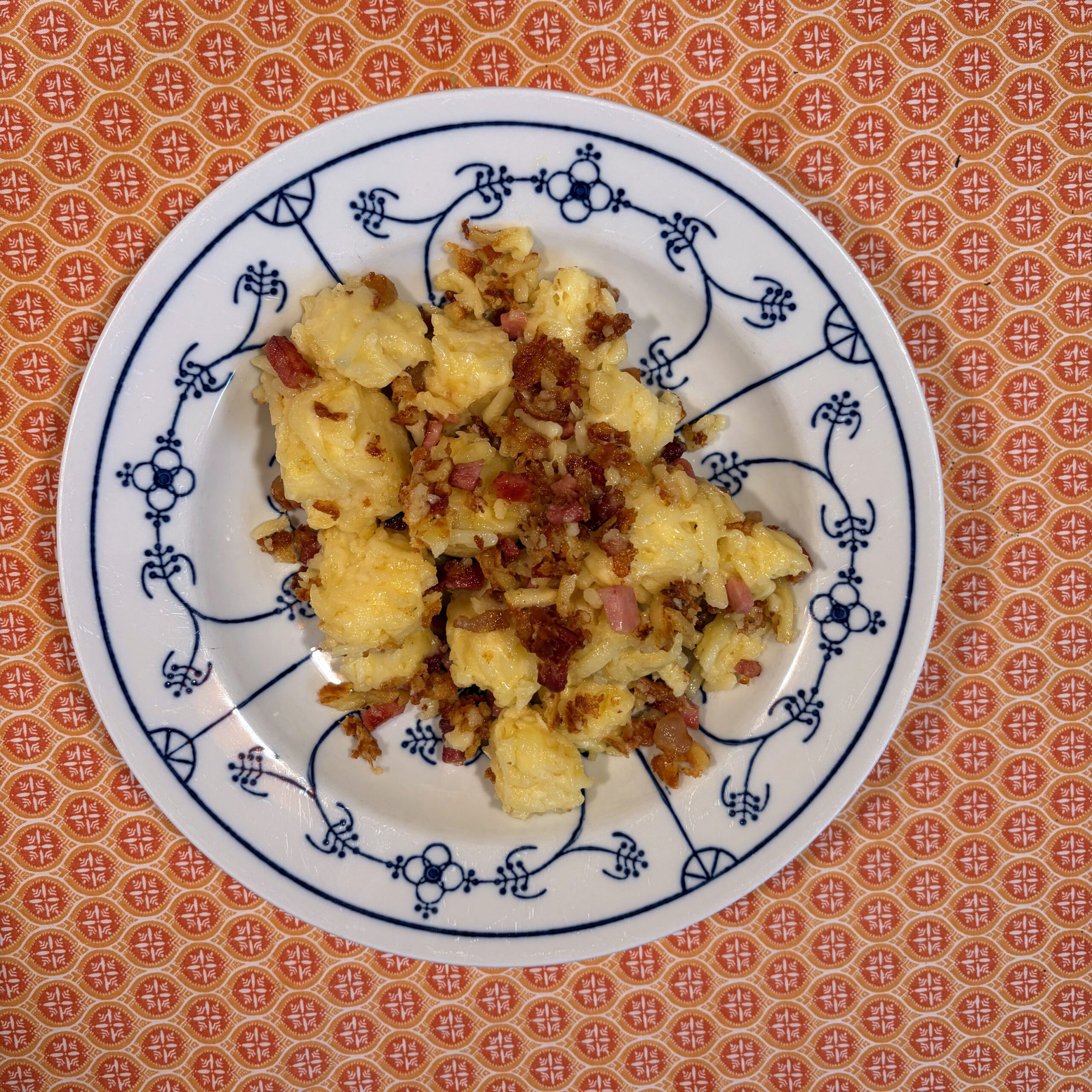 Aufgewärmte Käsespätzle mit gebräunten Röstzwiebeln und Speckwürfeln auf einem weiß-blauem Teller mit floralem Muster, serviert auf einem orangefarbenen gemusterten Untergrund.