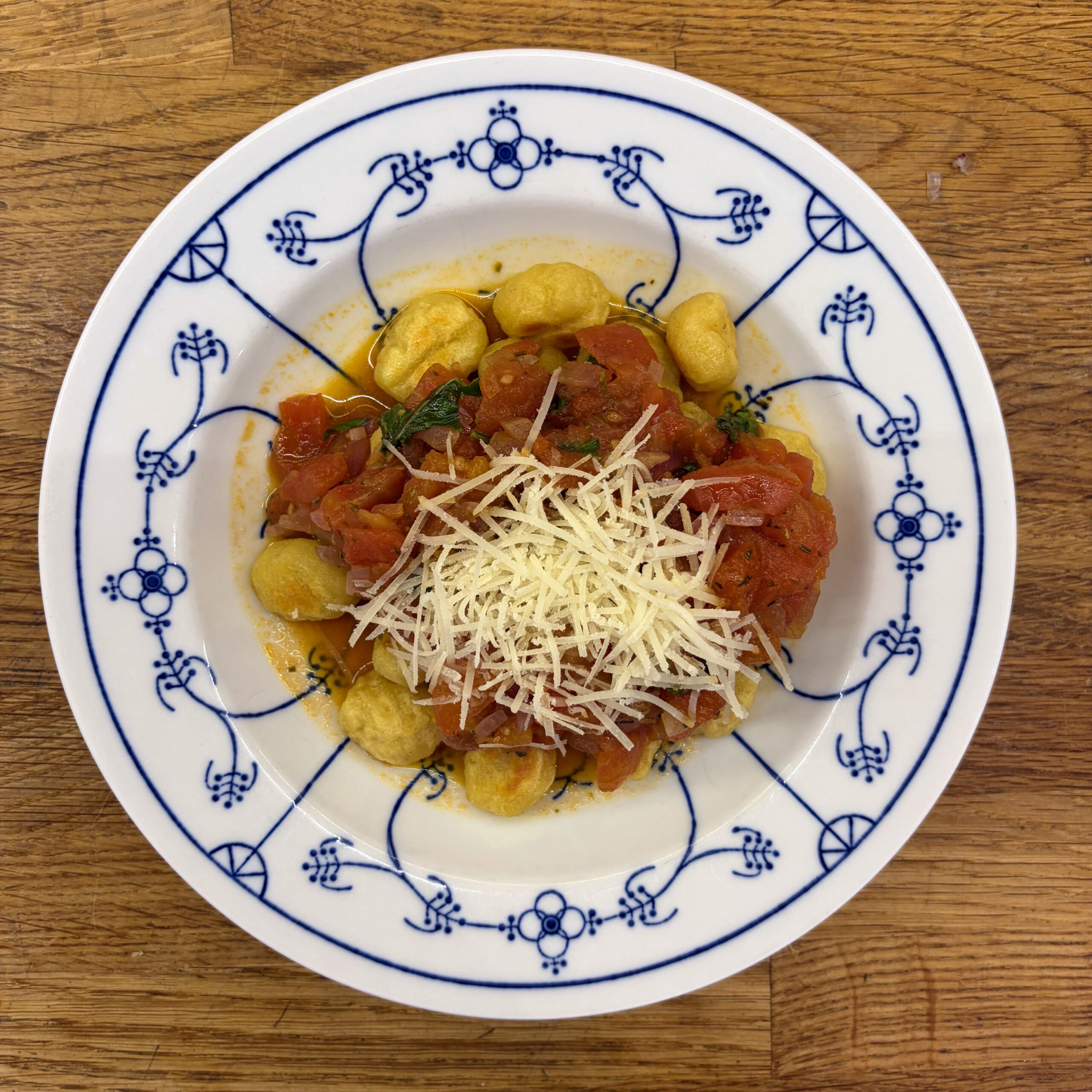 Gebackene Gnocchi mit Basilikum-Tomatensoße und geriebenem Parmesan auf einem weiß-blau gemusterten Teller, serviert auf einem Holztisch.