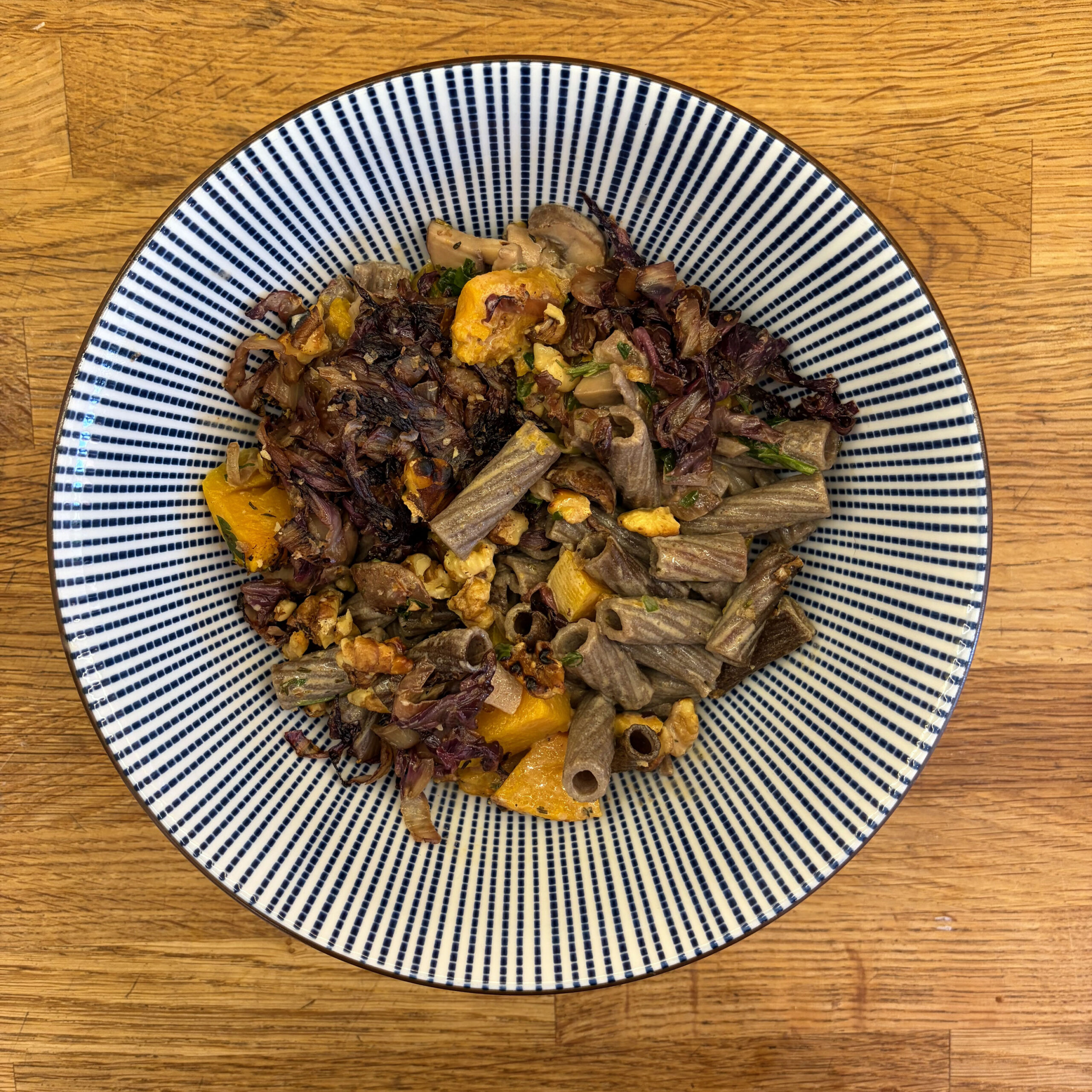 Ein Teller mit Buchweizen-Rigatoni-Auflauf, belegt mit gebratenen Champignons, geröstetem Kürbis und mariniertem Radicchio, serviert auf einem blau-weiß gestreiften Teller auf einem Holztisch.