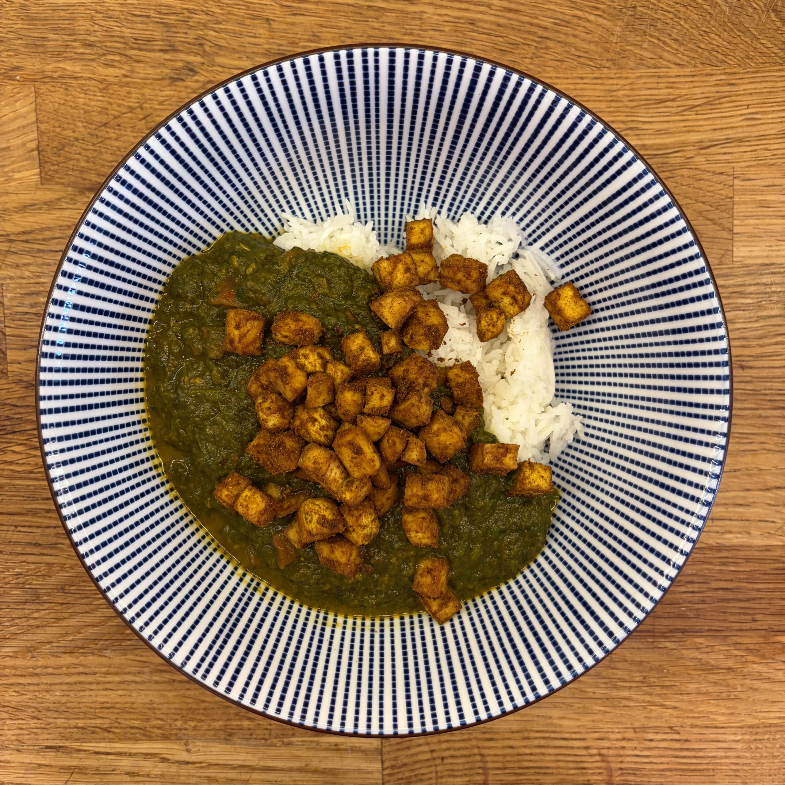 Teller mit Palak Tofu: grüne Spinatsauce mit gewürzten, goldbraun gebackenen Tofuwürfeln und weißem Basmatireis auf einem blau-weiß gestreiften Teller.