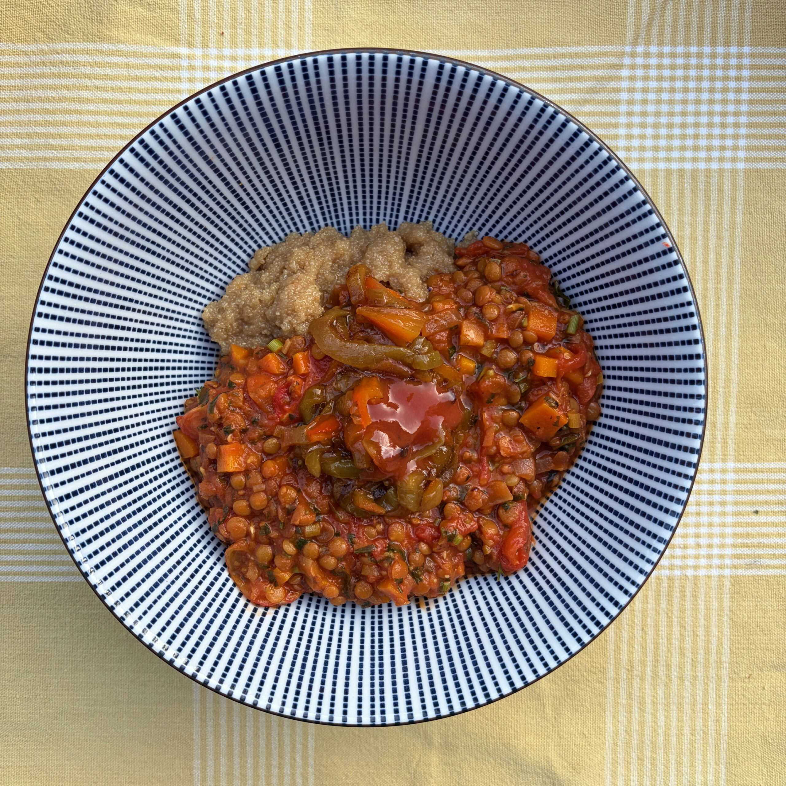 Draufsicht auf einen weiß-blau gestreiften Teller mit Amaranth-Polenta und Linsensoße: Eine tomatenbasierte Soße mit Berglinsen, Karotten, Sellerie und Zwiebeln bedeckt den größten Teil des Tellers, in der Mitte ein kleiner Schöpfer cremiger Amaranth-Polenta.
