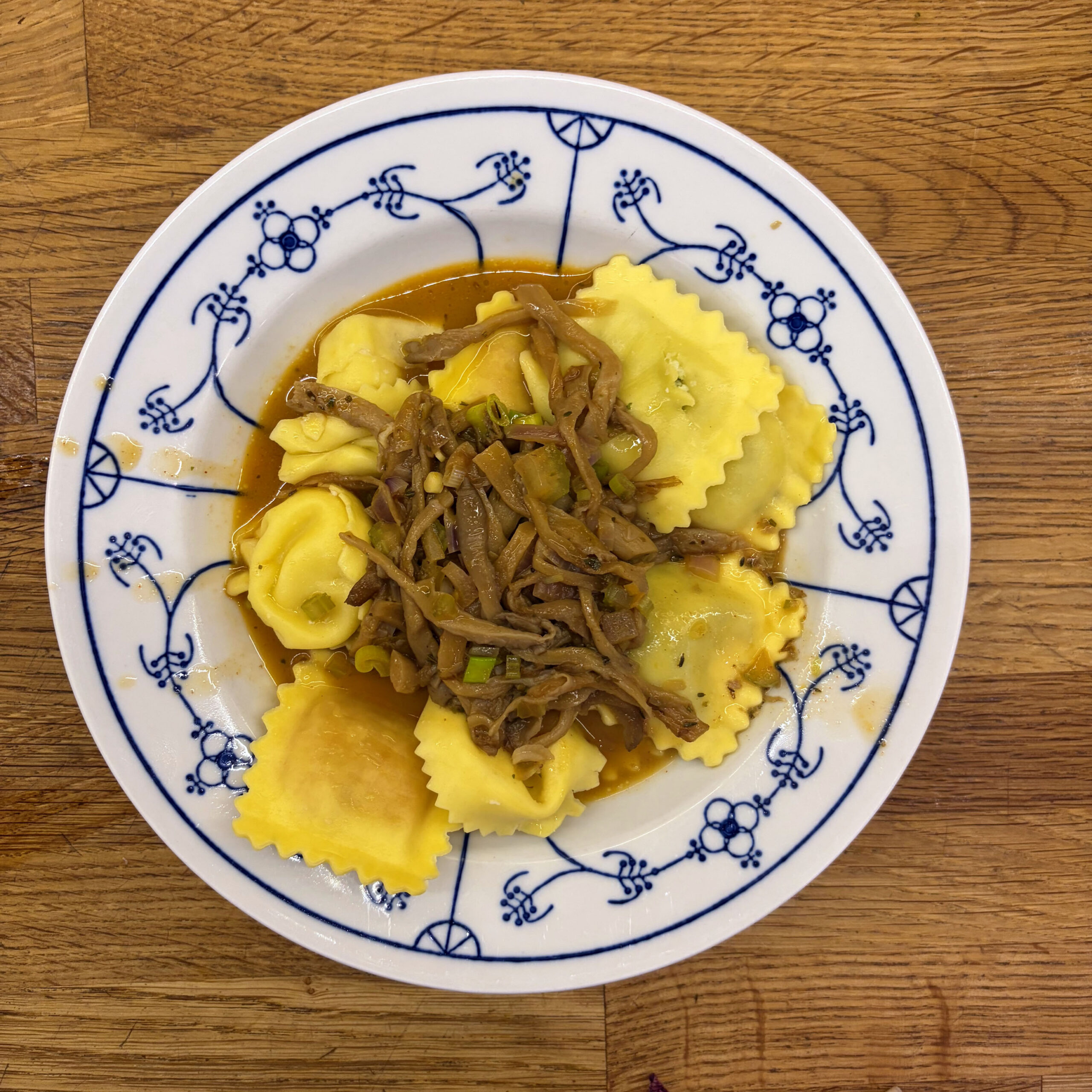 Dreierlei gefüllte Tortellini mit Tomate-Ricotta, Spinat-Ricotta und Parmesan, angerichtet mit gebratenen Pfifferlingen und Zwiebeln in Hafersahne-Soße auf einem weiß-blau gemusterten Teller.