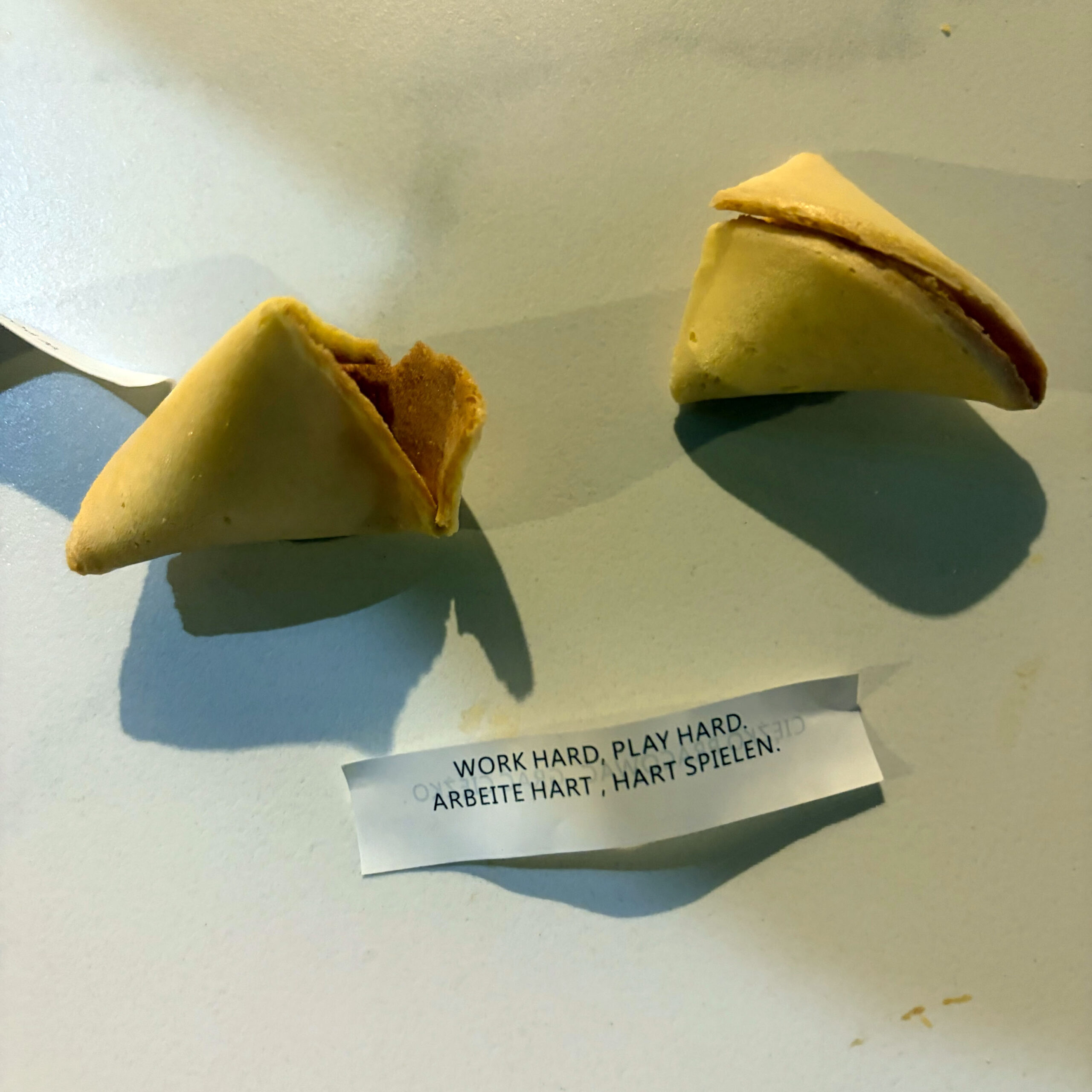 Zwei Glückskekse auf einer hellen Oberfläche, einer davon aufgebrochen. Der herausgezogene Zettel trägt die Aufschrift: „Work hard, play hard. / Arbeite hart, hart spielen."