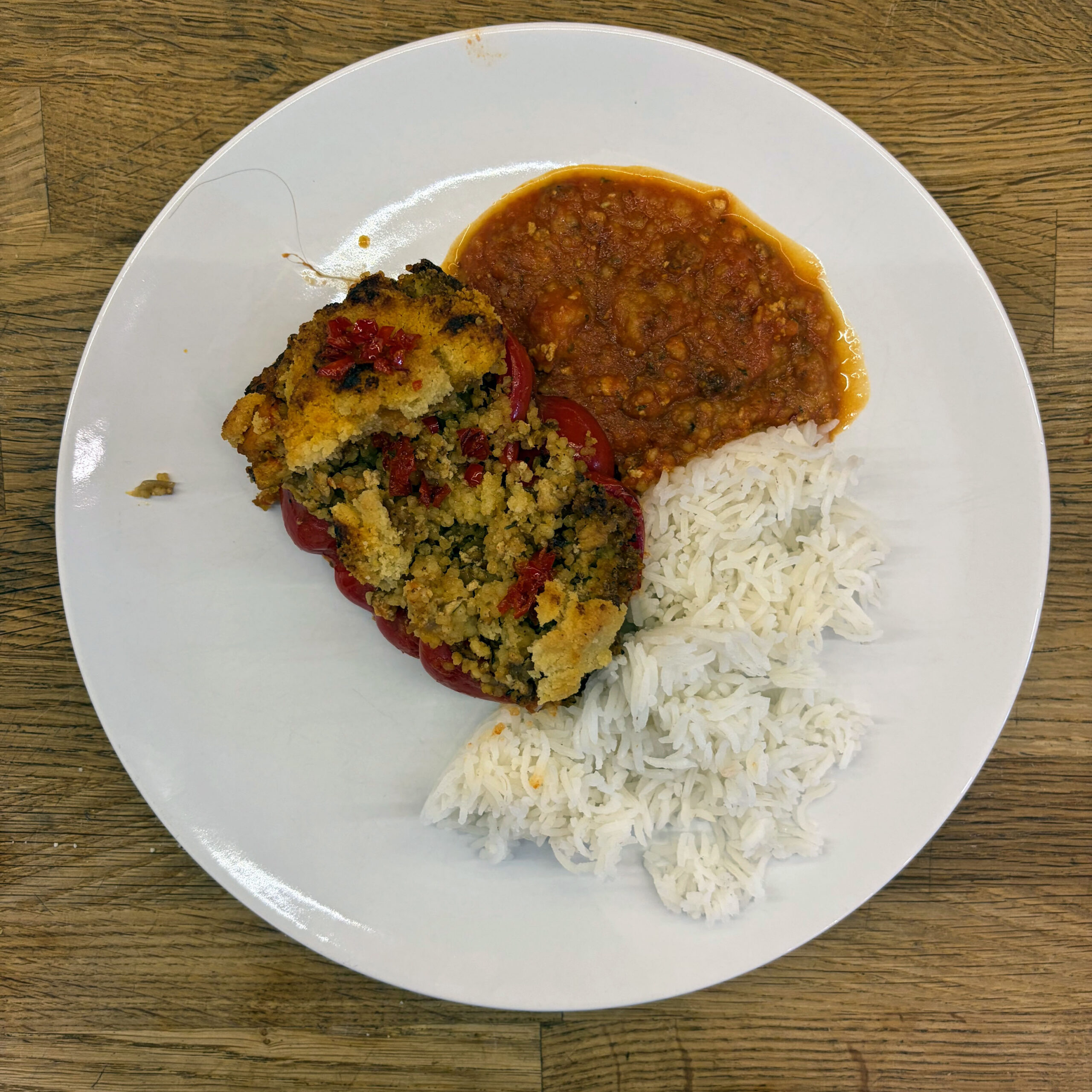 Eine halbe rote Paprika, gefüllt mit einer Hirse-Tofu-Hack-Mischung und gerösteten Semmelbröseln, serviert mit weißem Reis und einer stückigen Tomatensoße auf einem weißen Teller.