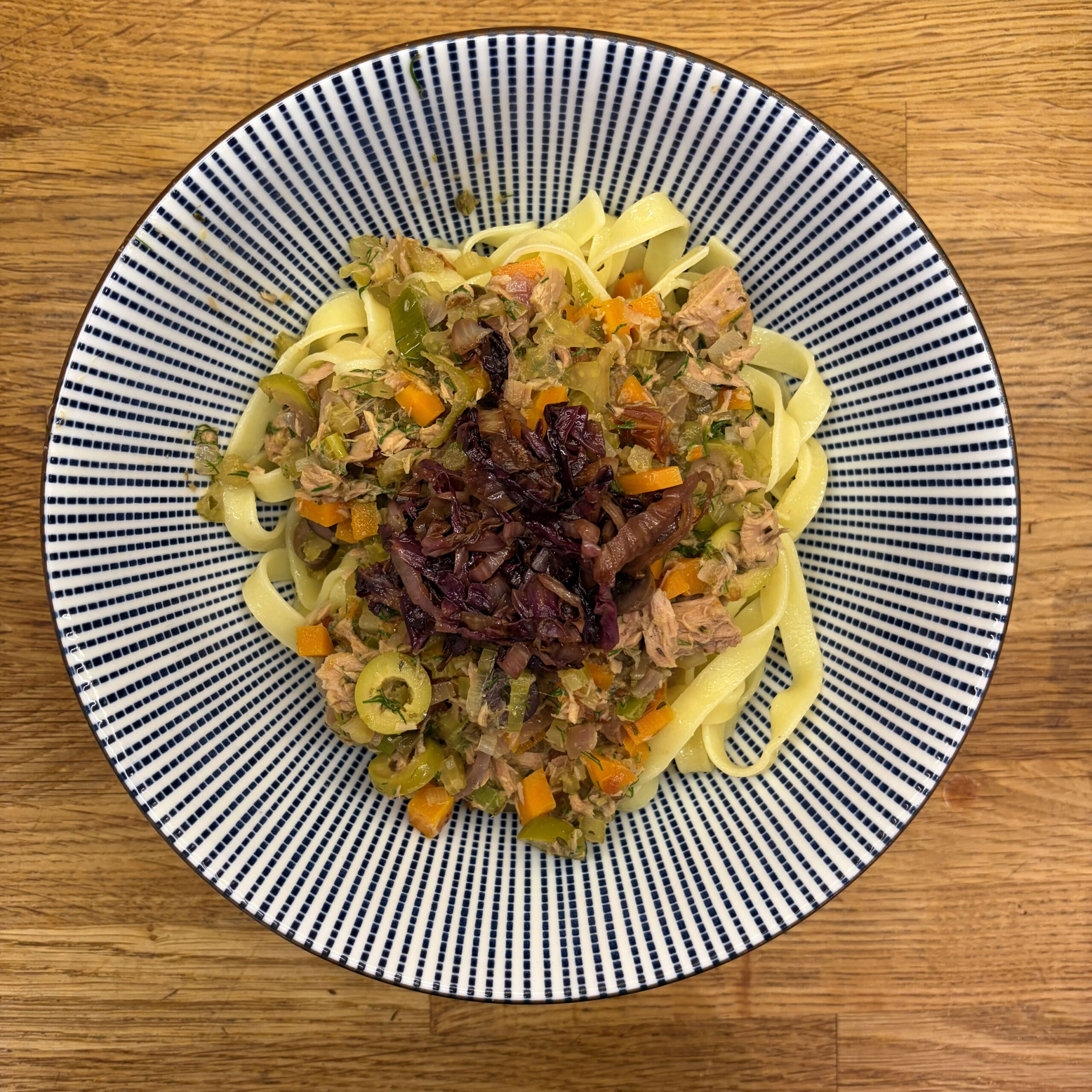 Tagliatelle mit Thunfischsoße aus Zwiebeln, Sellerie, Möhren, Oliven, getrockneten Tomaten und Kapern, garniert mit gebratenem Radicchio in Balsamico-Marinade, serviert in einer blau-weiß gestreiften Schüssel auf einem Holztisch.