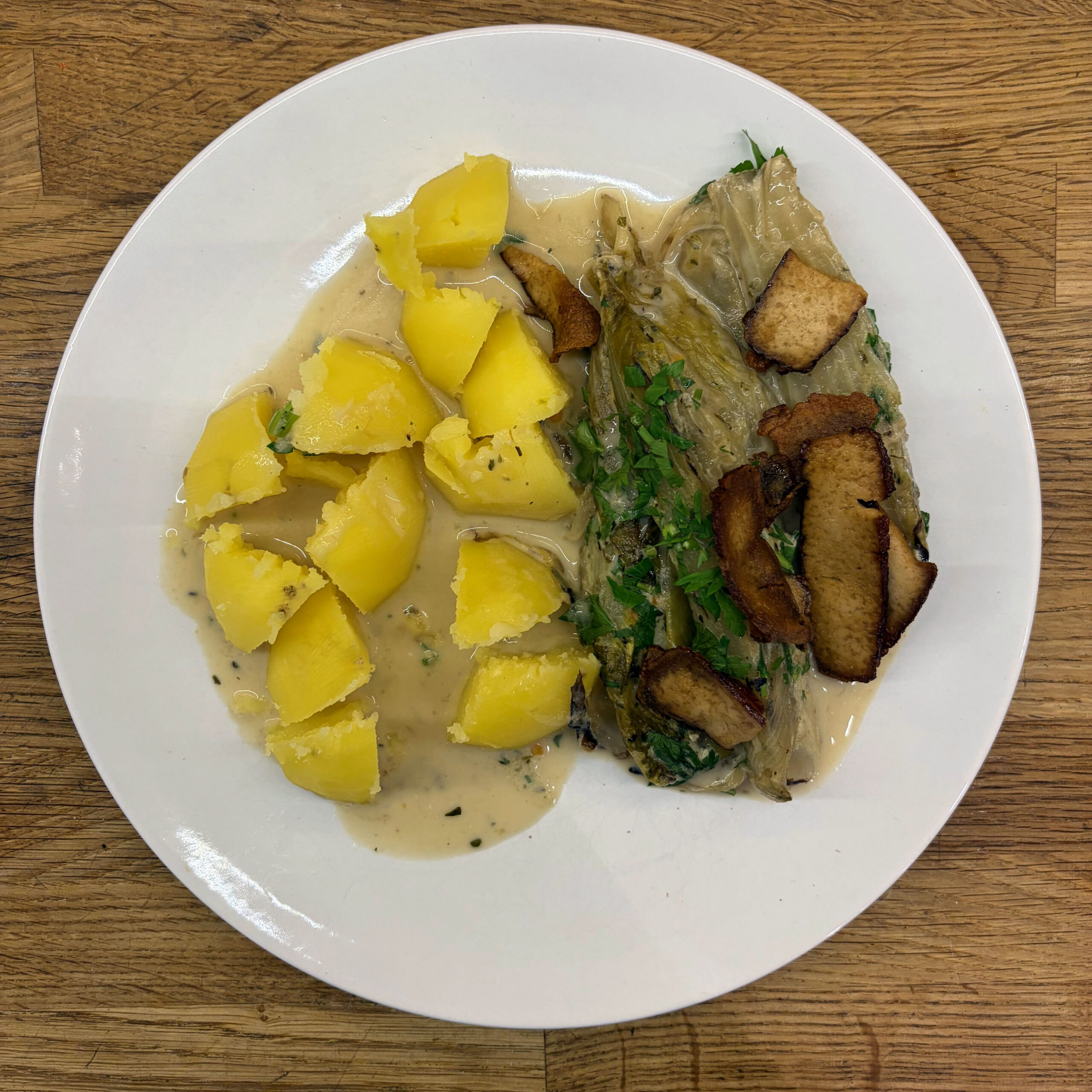 Teller mit gebratenem Chicorée in Mandelsahne-Sauce mit gehackter Petersilie, gekochten Salzkartoffeln und knusprig gebratenen Räuchertofu-Scheiben, serviert auf weißem Teller.