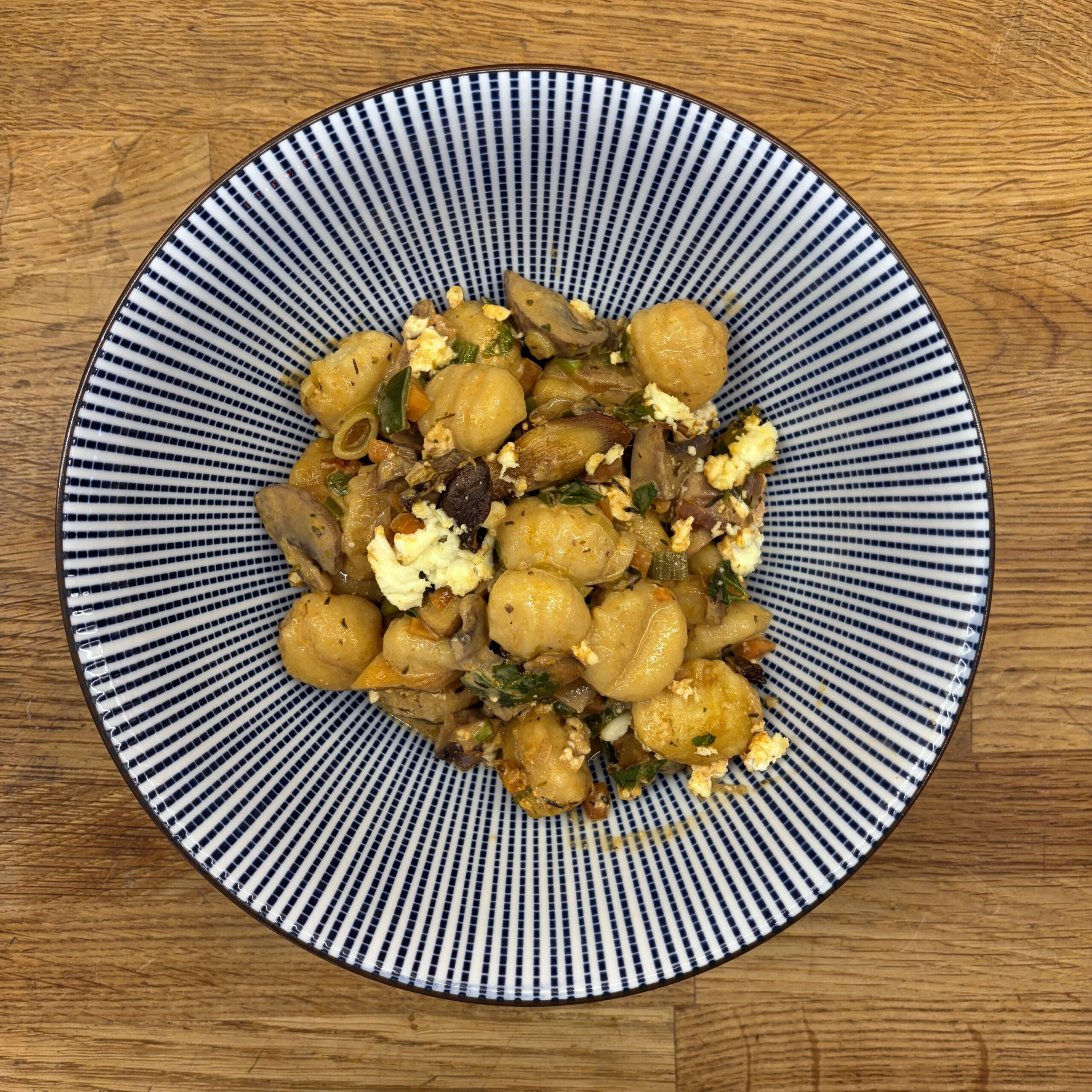 Gebratene Gnocchi mit Champignons, Kräuterseitlingen, Möhren und Lauchzwiebeln in Sahnesoße, garniert mit zerbröckeltem Feta, serviert auf einem blau-weiß gestreiften Teller auf einem Holzuntergrund.
