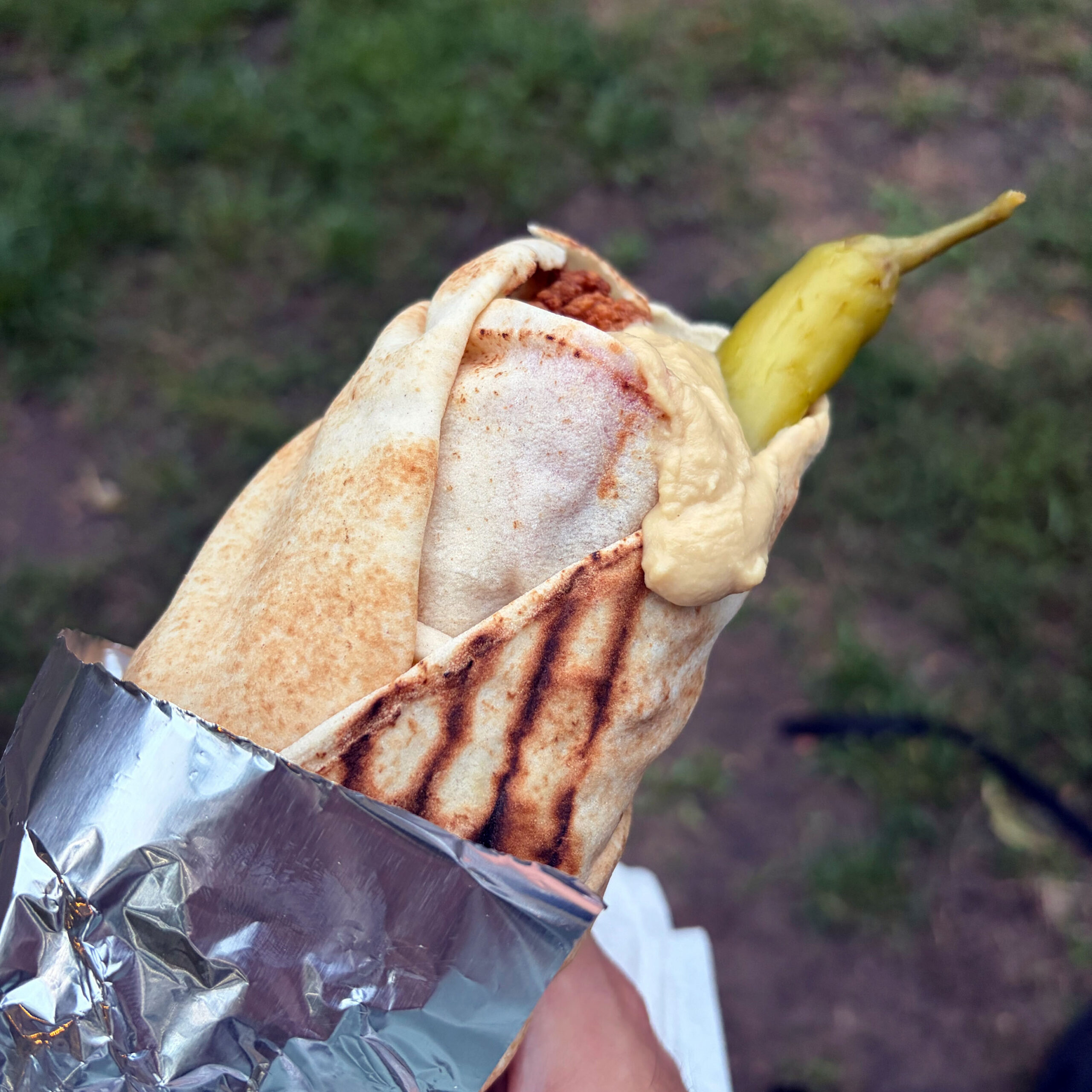 Gegrilltes Halloumi-Makali-Wrap in Fladenbrot, gefaltet und in Alufolie gewickelt, mit Hummus und einer eingelegten Pepperoni on top.