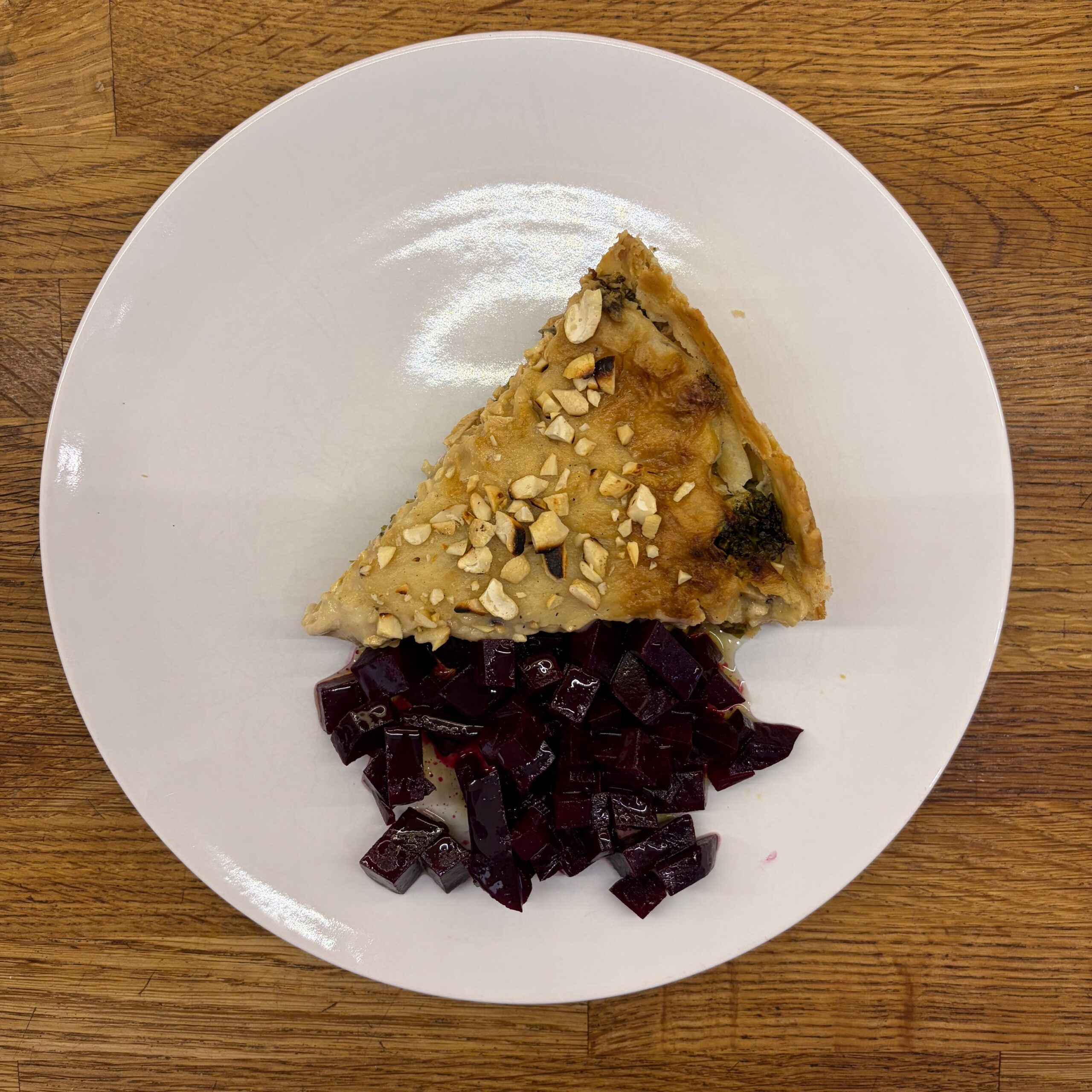 Ein Stück Brokkoli-Tofu-Tarte mit goldbrauner Mürbeteigkruste und gehackten Cashewkernen auf einer weißen Platte, begleitet von gewürfelter, in Balsamico marinierter Roter Bete.