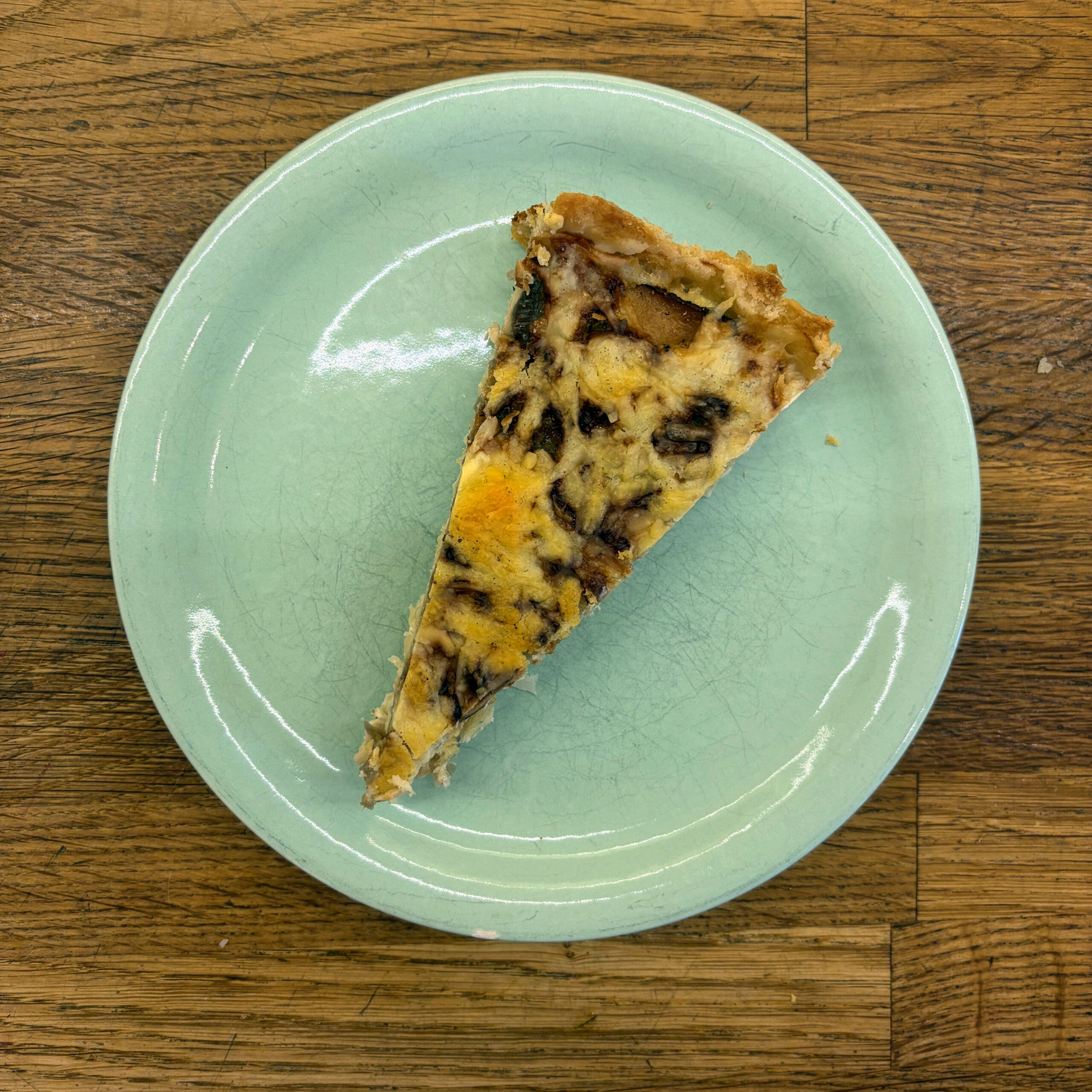 Ein Stück Sauerkraut-Tarte mit sichtbaren Füllungsschichten aus Sauerkraut, karamellisierten Zwiebeln, Räuchertofu und geschmolzenem Bergkäse in einem Mürbeteigboden, serviert auf einem mintgrünen Keramikteller auf einem Holztisch.