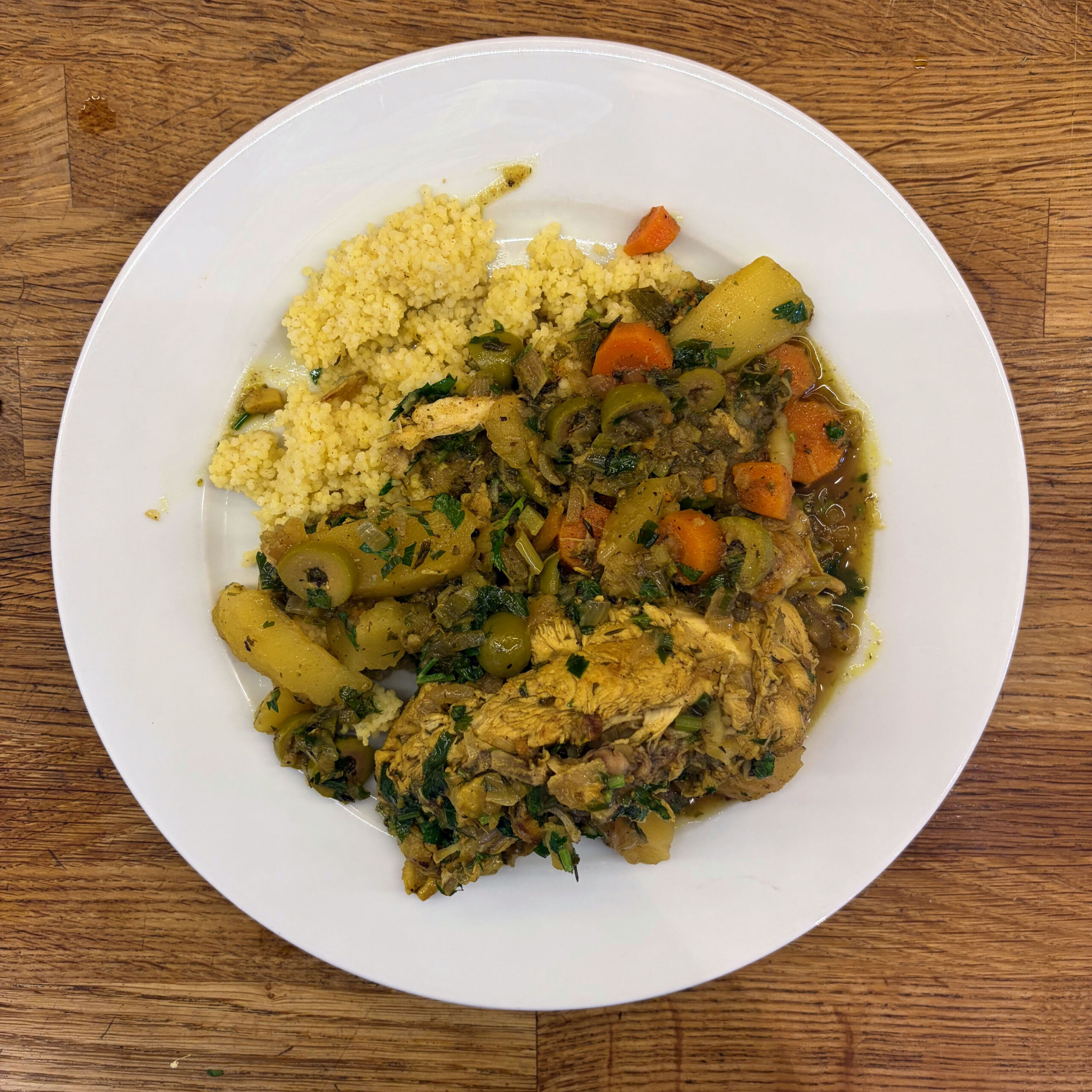 Weißer Teller mit Hähnchen-Tajine mit Salzzitronen, grünen Oliven, Kartoffeln und Möhren in würziger Sauce, serviert mit Hirse, auf einer Holzunterlage.