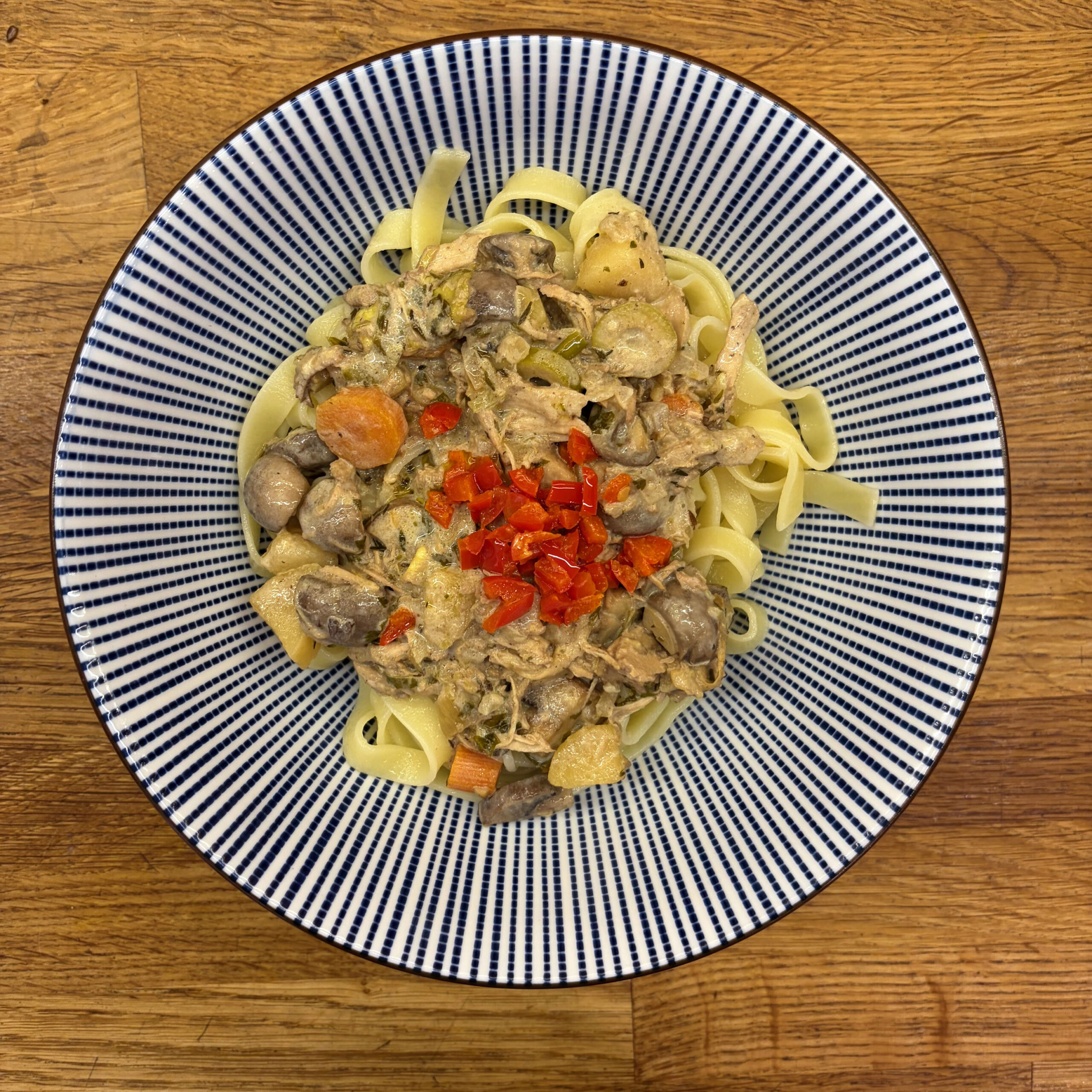 Tagliatelle auf einem blau-weiß gestreiften Teller, belegt mit einer cremigen Champignonsoße aus Restesoße und Mandelsahne, garniert mit gewürfelten roten Paprikastücken.