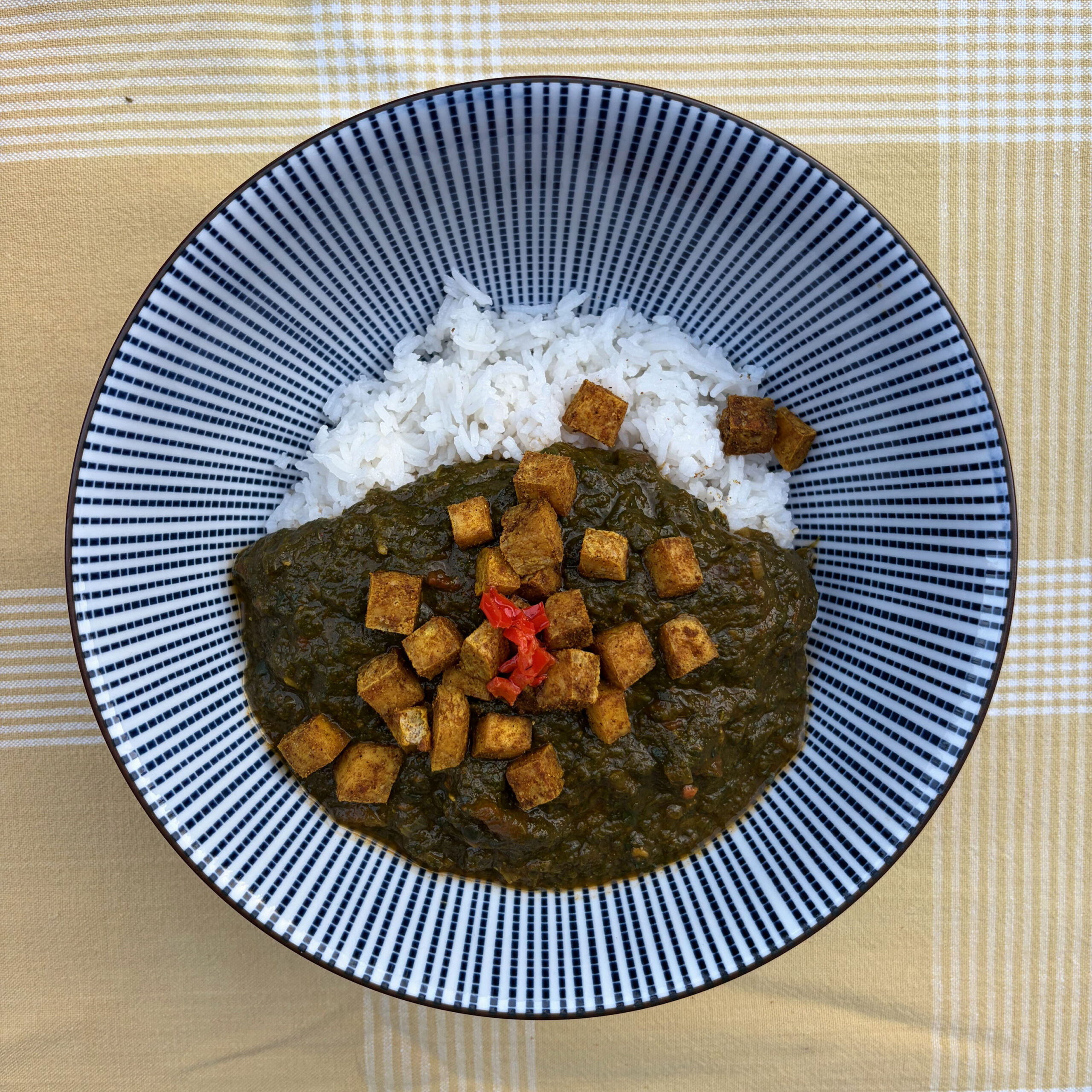 Palak Tofu in einer blau-weiß gestreiften Schüssel: dunkles Spinatcurry mit gewürzten, gebackenen Tofuwürfeln und einem roten Habanero-Stück in der Mitte, daneben weißer Basmatireis.