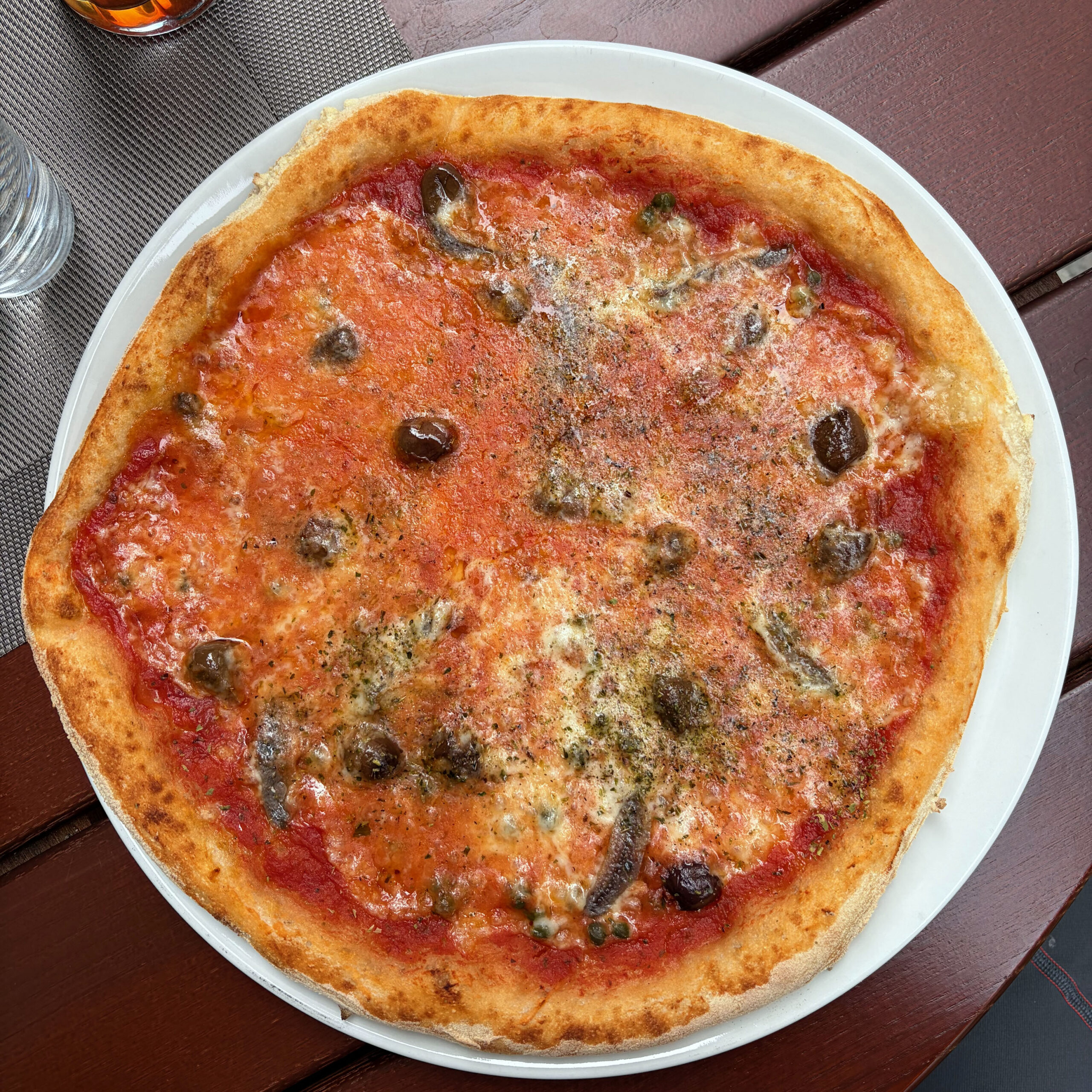 Pizza Napoletana mit Tomatensauce, Sardellen, schwarzen Oliven und Kapern auf weißem Teller, serviert auf einem Holztisch in der Rastkogelhütte.