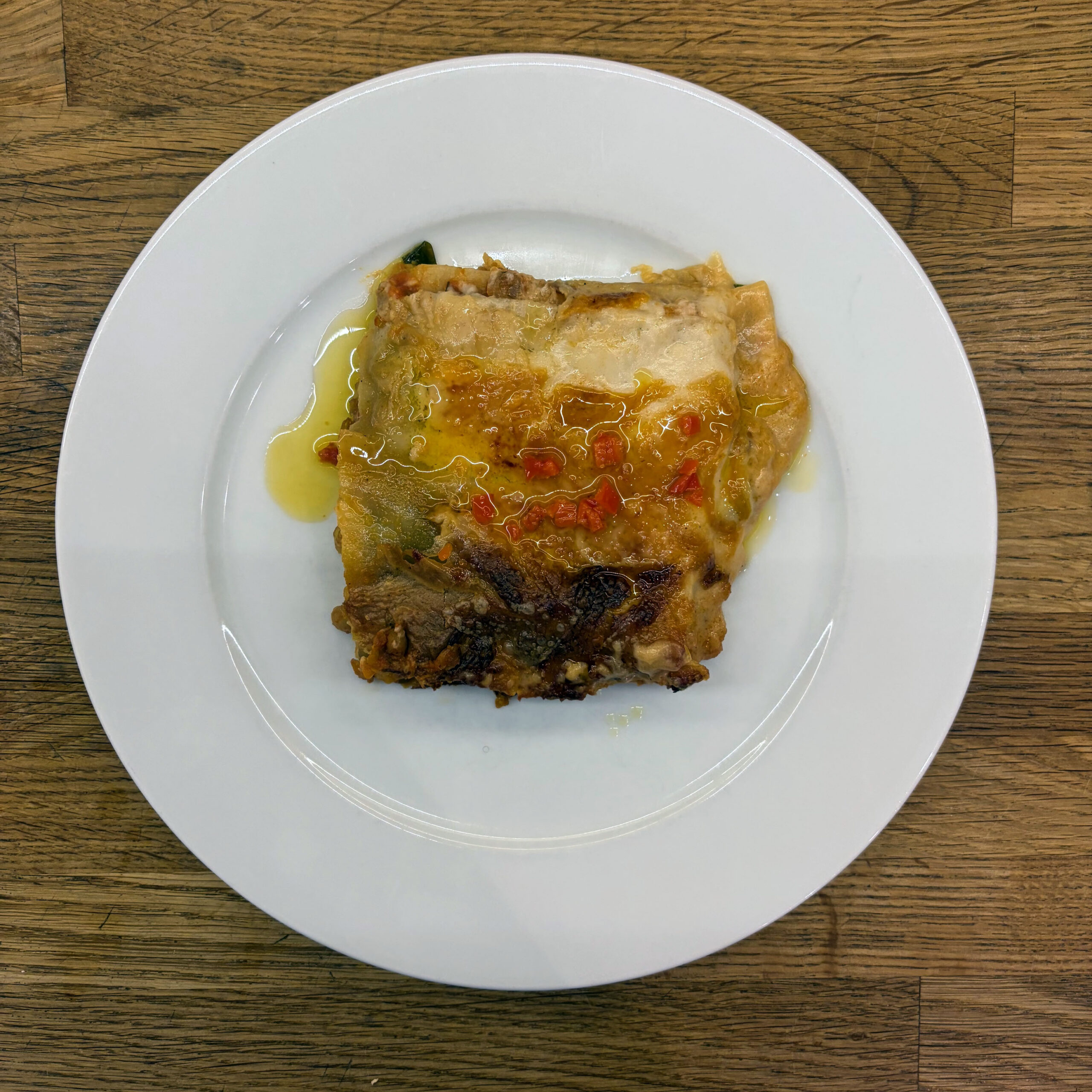 Ein Stück gebackene Pizza Rustica mit Sardellen, Kapern und Oliven auf einem weißen Teller, serviert mit etwas Olivenöl, auf einem Holztisch.