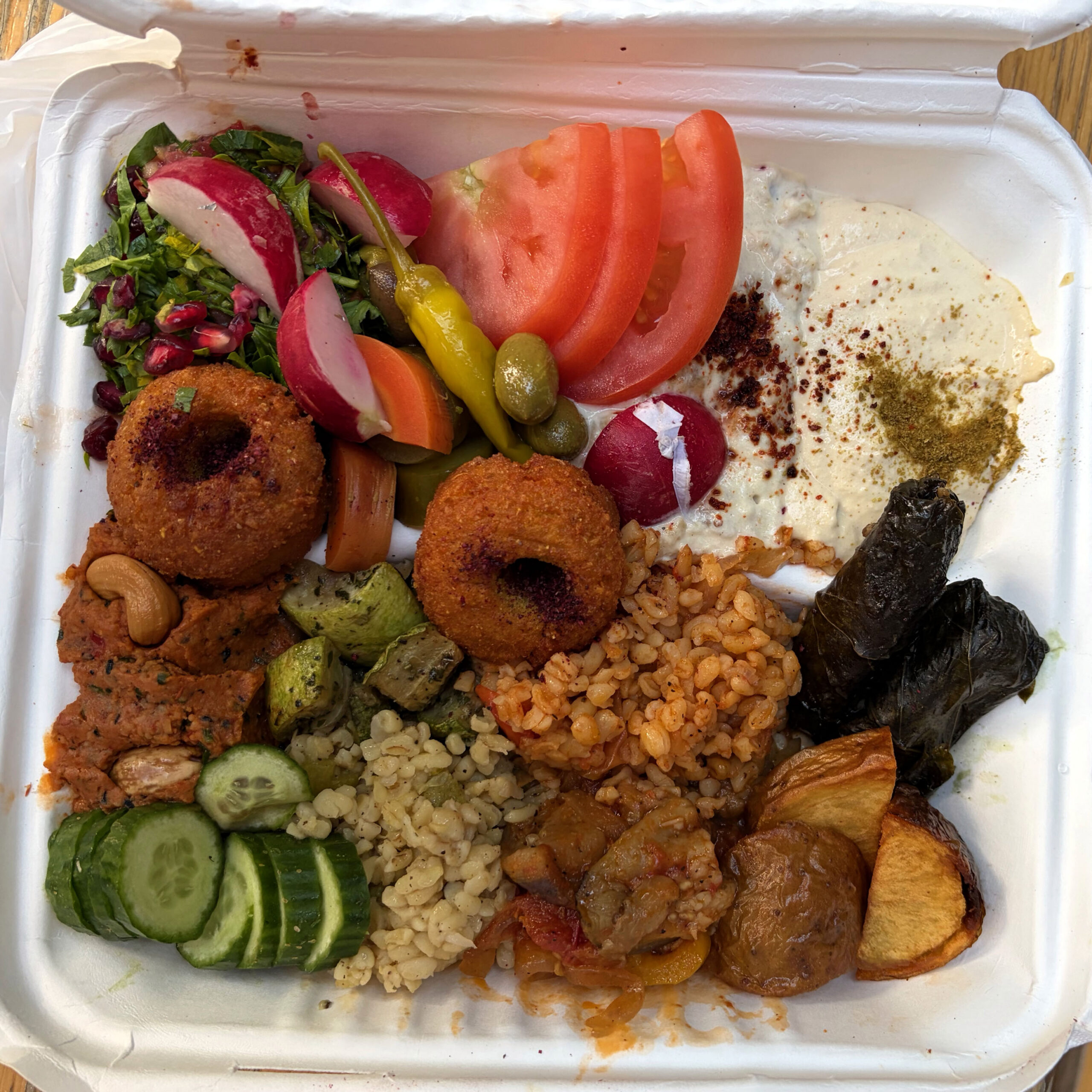 Eine Styroporbox gefüllt mit einer Auswahl veganer Mezze: Kibbeh (frittierte Bulgurbällchen), Hummus mit Gewürzen, gefüllte Weinblätter (Dolmades), Bulgursalat, geröstete Kartoffeln, gegrillte Zucchini, Tabbouleh mit Granatapfelkernen, frische Tomaten- und Radieschenscheiben, eingelegte Peperoni und Kapern sowie Gurkenscheiben.