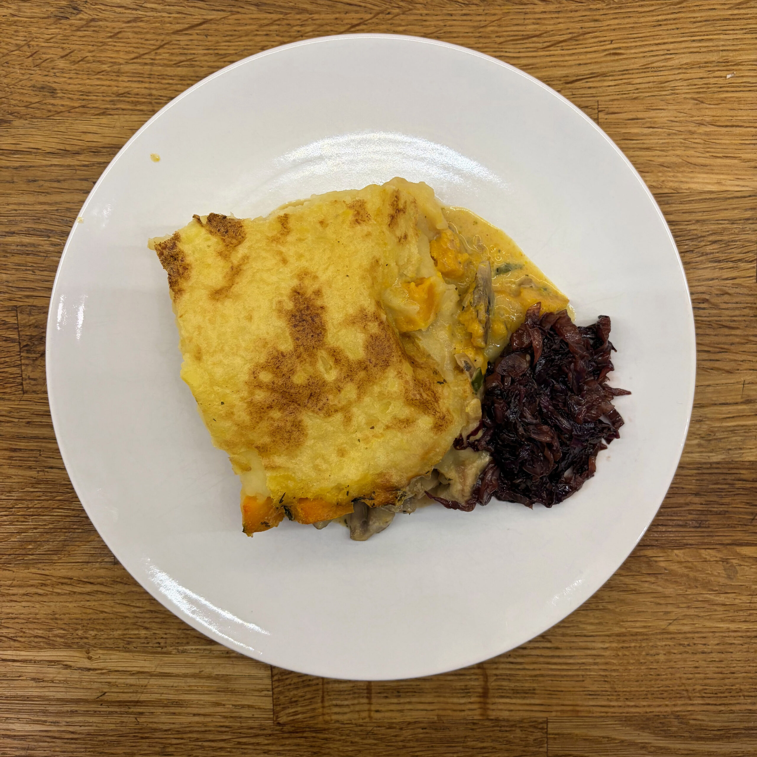 Ein Stück veganer Shepherd's Pie mit goldbraun gebackenem Kartoffelpüree auf einer Schicht Champignon-Mandelsahne und Hokkaido-Kürbis, serviert mit mariniertem Radicchio, auf einem weißen Teller.
