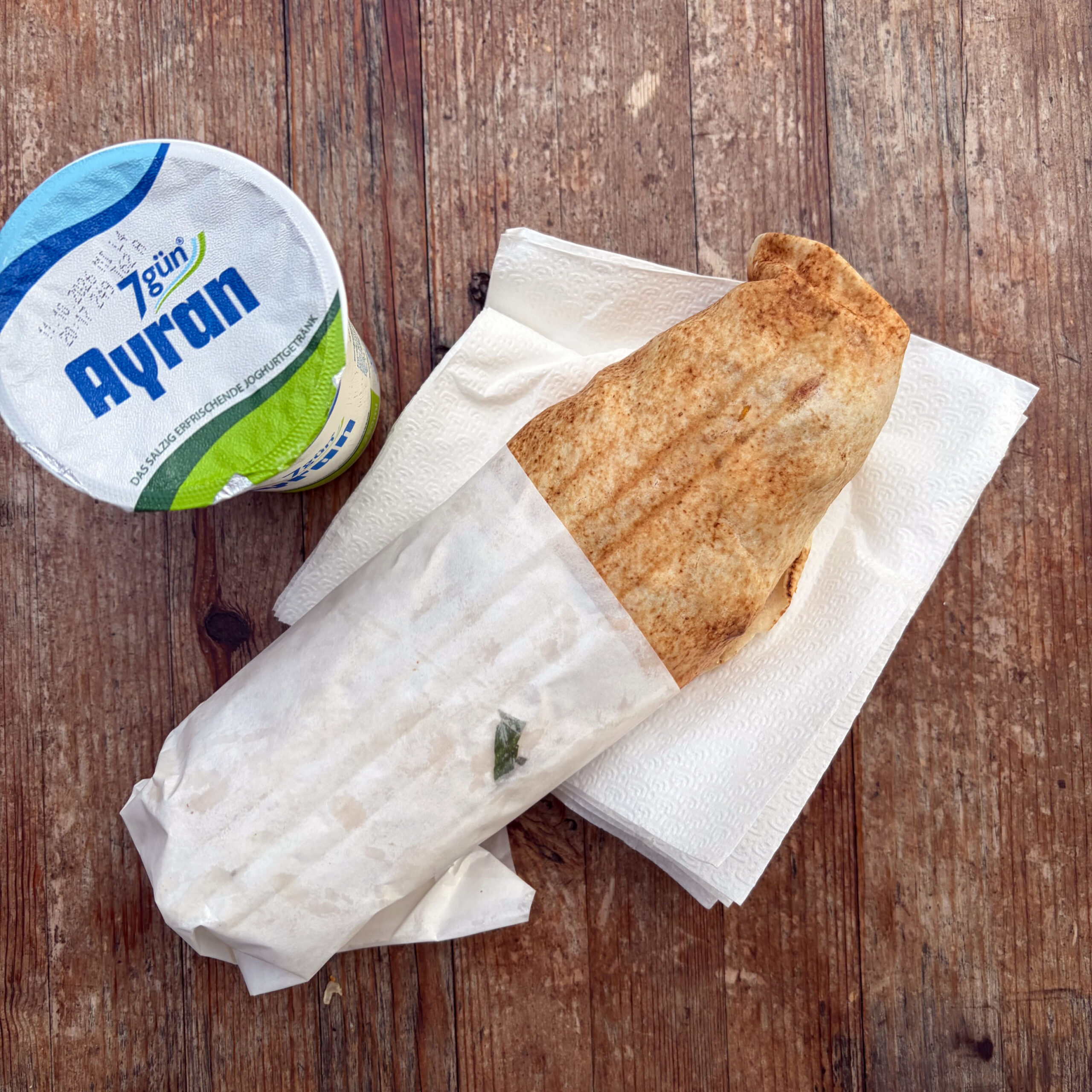 Gegrilltes Falafel-Sandwich in Fladenbrot, in Papier eingewickelt auf Servietten liegend, daneben ein Becher Ayran von 7gün, auf einem Holztisch.