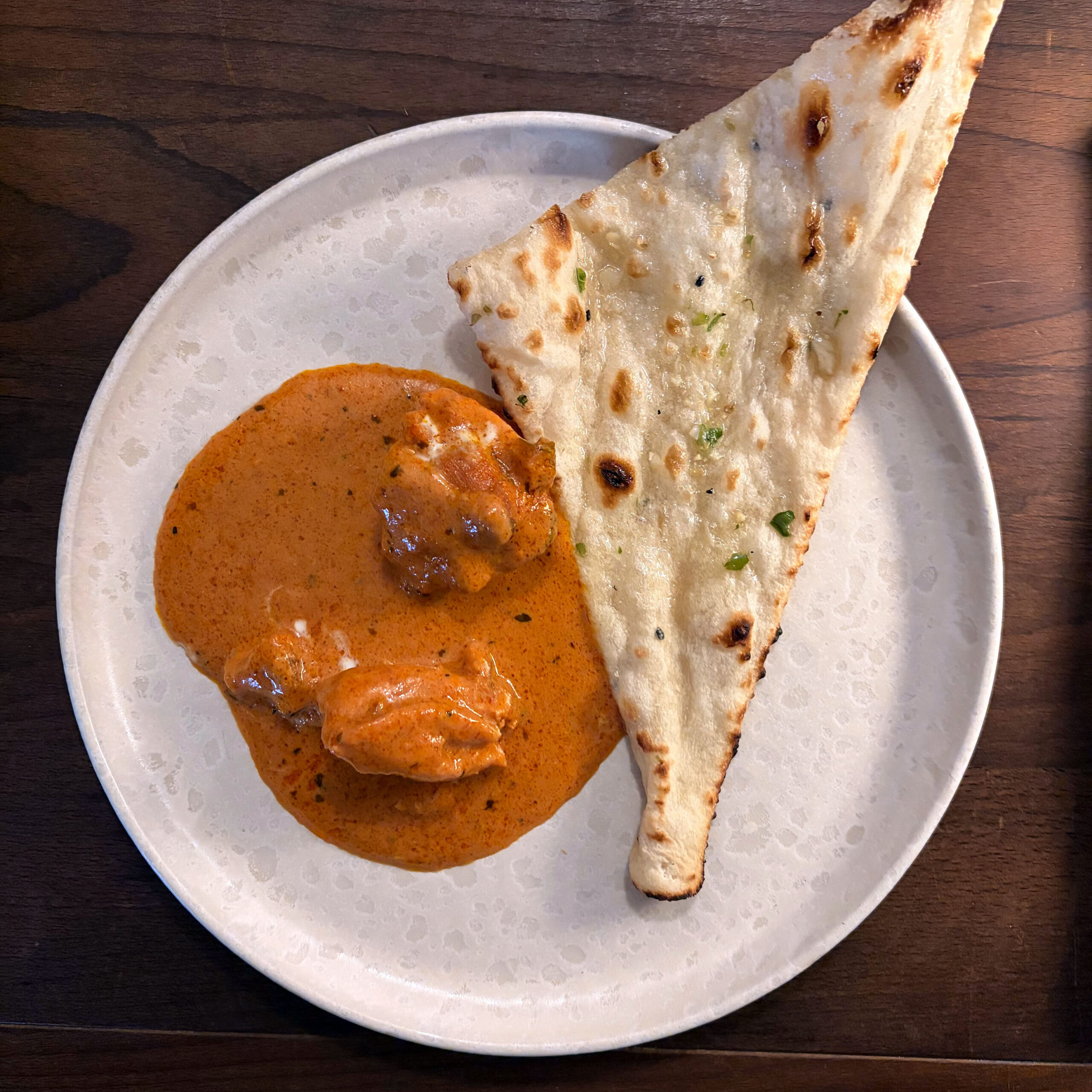 Butter Chicken in cremiger, orangeroter Currysauce mit Knoblauch-Chili-Naan auf weißem Teller, serviert auf einem Holztisch.