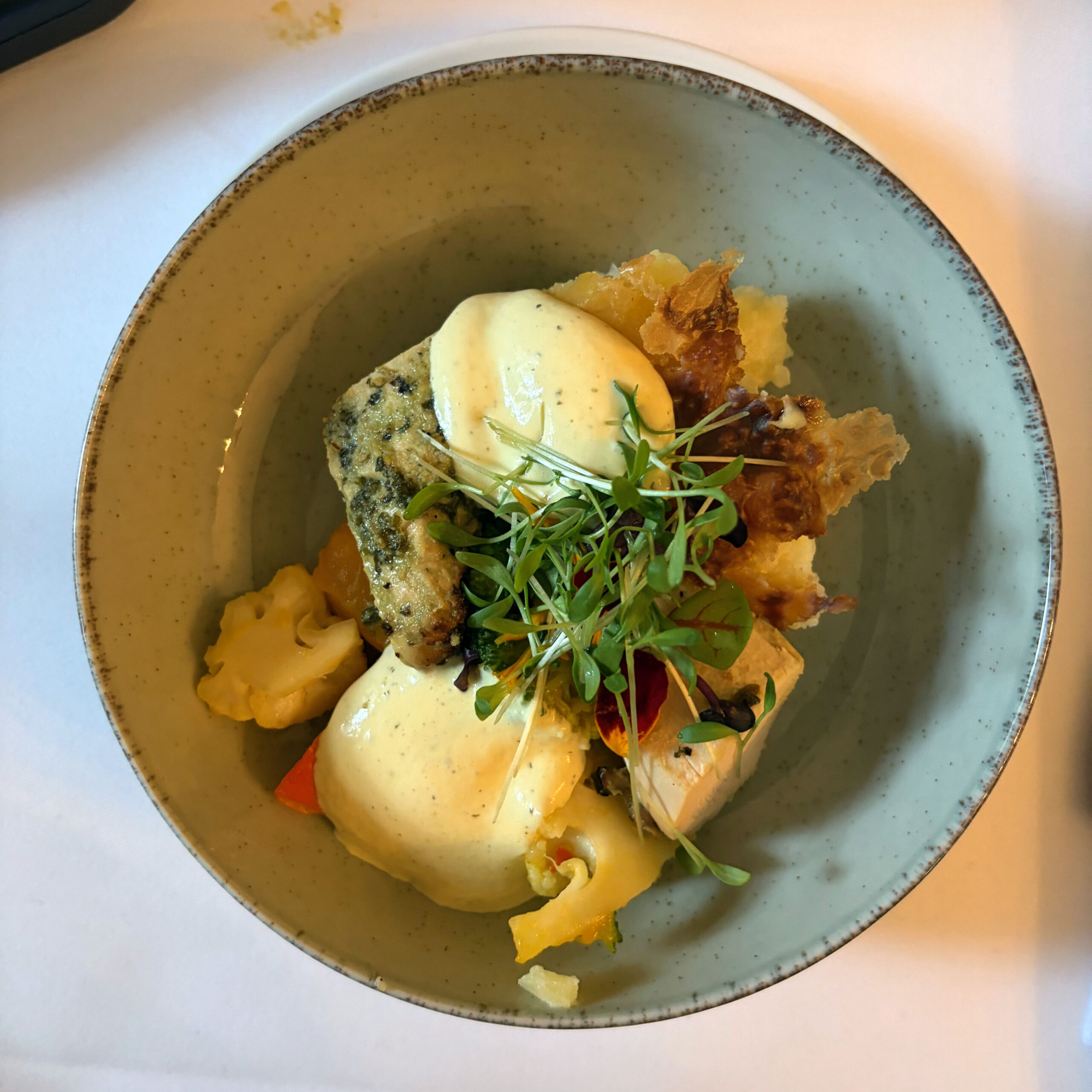 Fischfilet mit Kräuterkruste und cremiger Sauce auf einem Bett aus buntem Blumenkohl und Gemüse, garniert mit Microgreens und knuspriger Fischhaut, serviert in einer grauen Keramikschale.