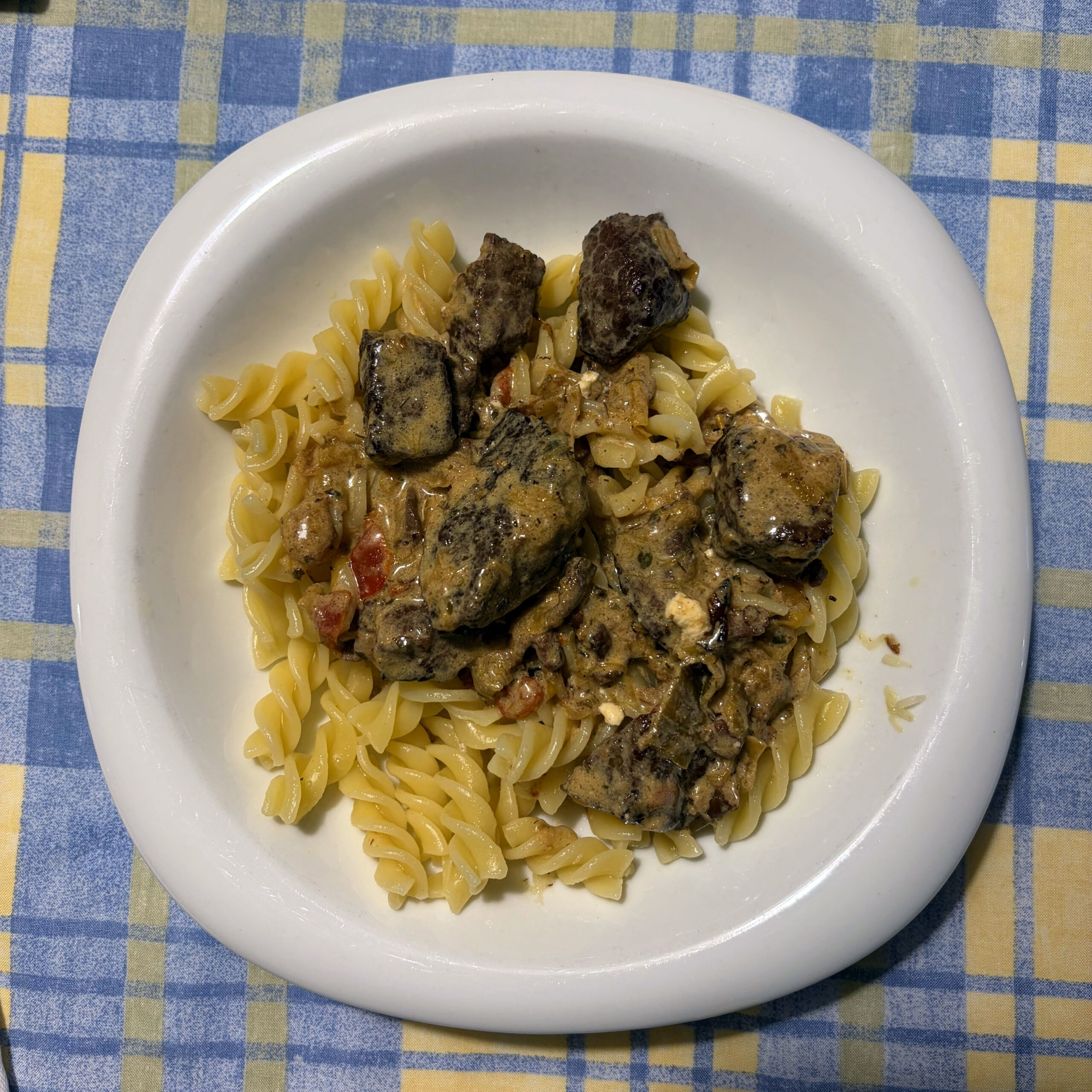 Teller mit Rindergulasch und Fusilli-Nudeln in würziger Soße mit Tomaten- und Zwiebelstücken, serviert auf blau-gelbem Karostoff.