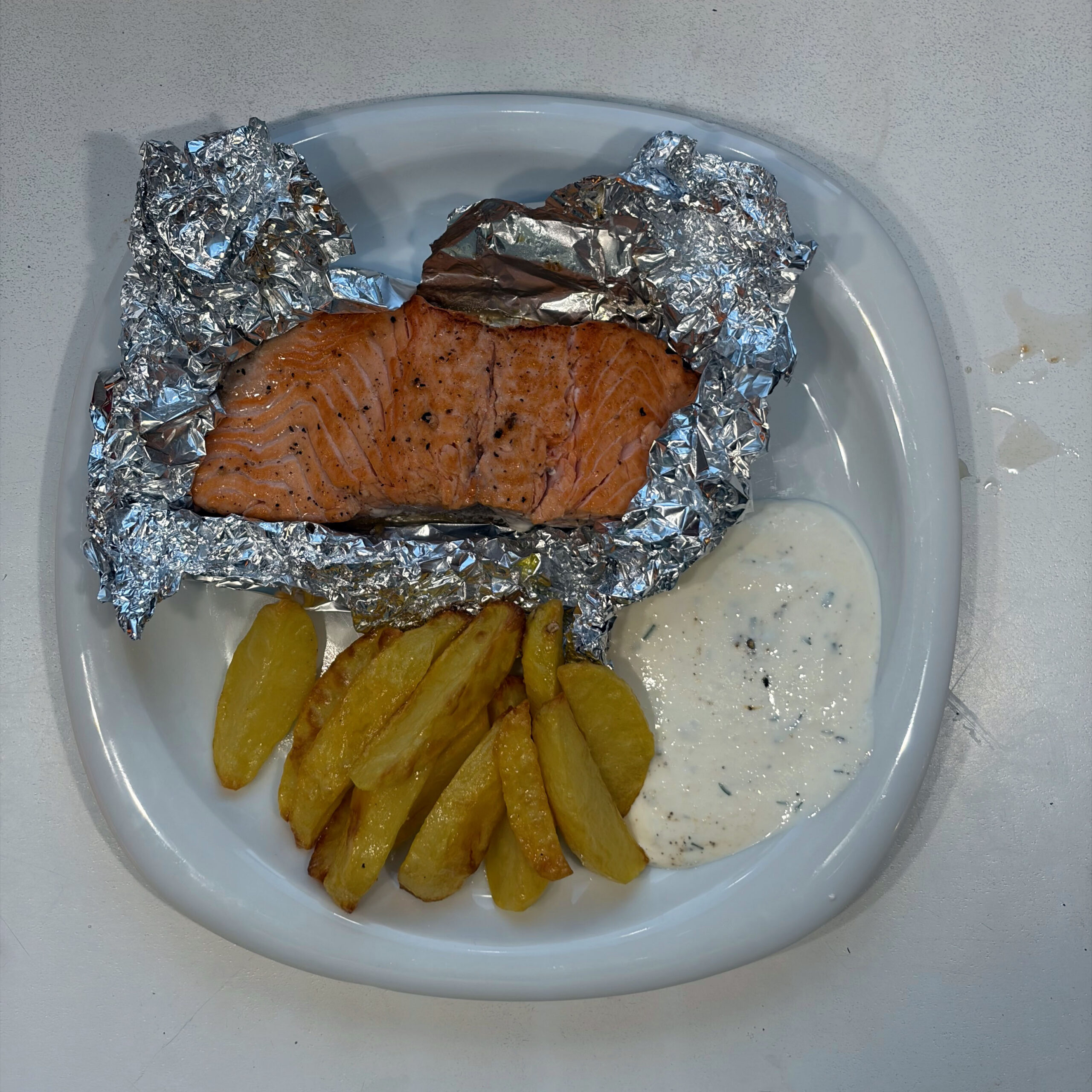 Lachsforellenfilet in geöffneter Alufolie auf einem weißen Teller, serviert mit goldbraunen Ofenkartoffelspalten und einem cremigen Joghurt-Dill-Dipp.