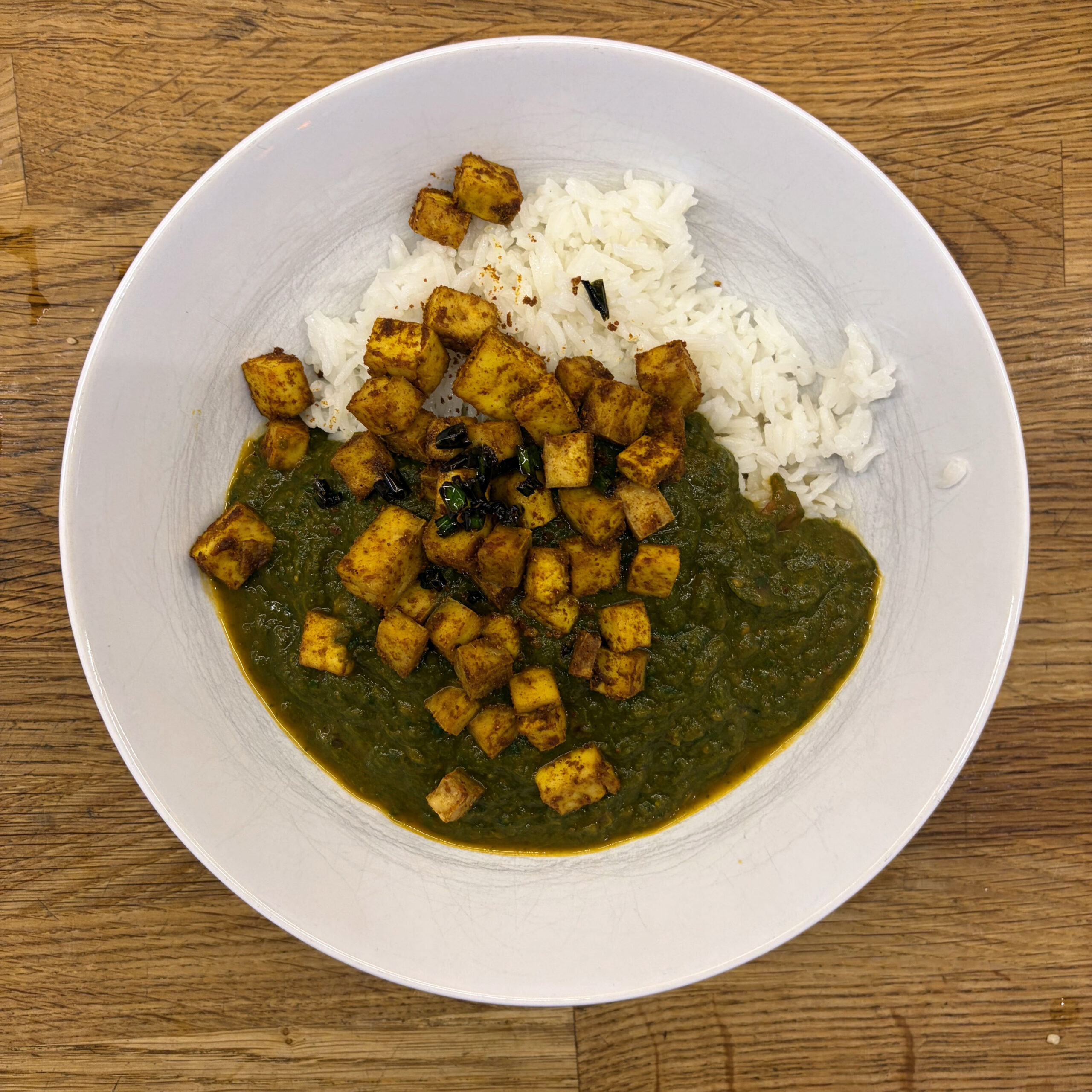 Eine weiße Schüssel mit Palak Tofu: dunkelgrüne Spinat-Tomaten-Sauce, belegt mit gewürzten, goldbraun gebackenen Tofuwürfeln und weißem Basmatireis, serviert auf einem Holzuntergrund.