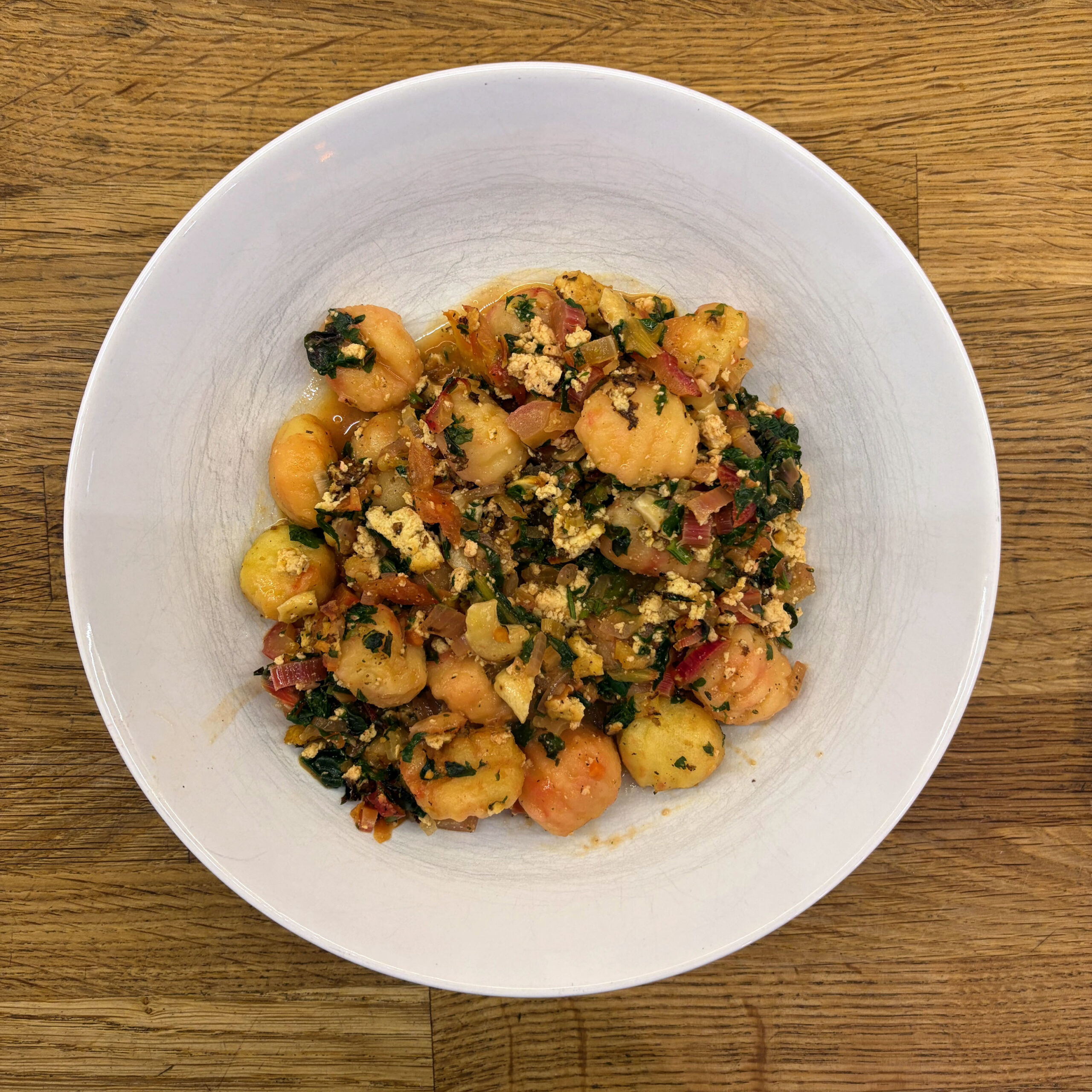 Weiße Schüssel mit gebackenen Gnocchi, gedünstetem Mangold, zerbröseltem Tofu und Gemüsesoße aus Tomaten und Zwiebeln auf einem Holztisch.