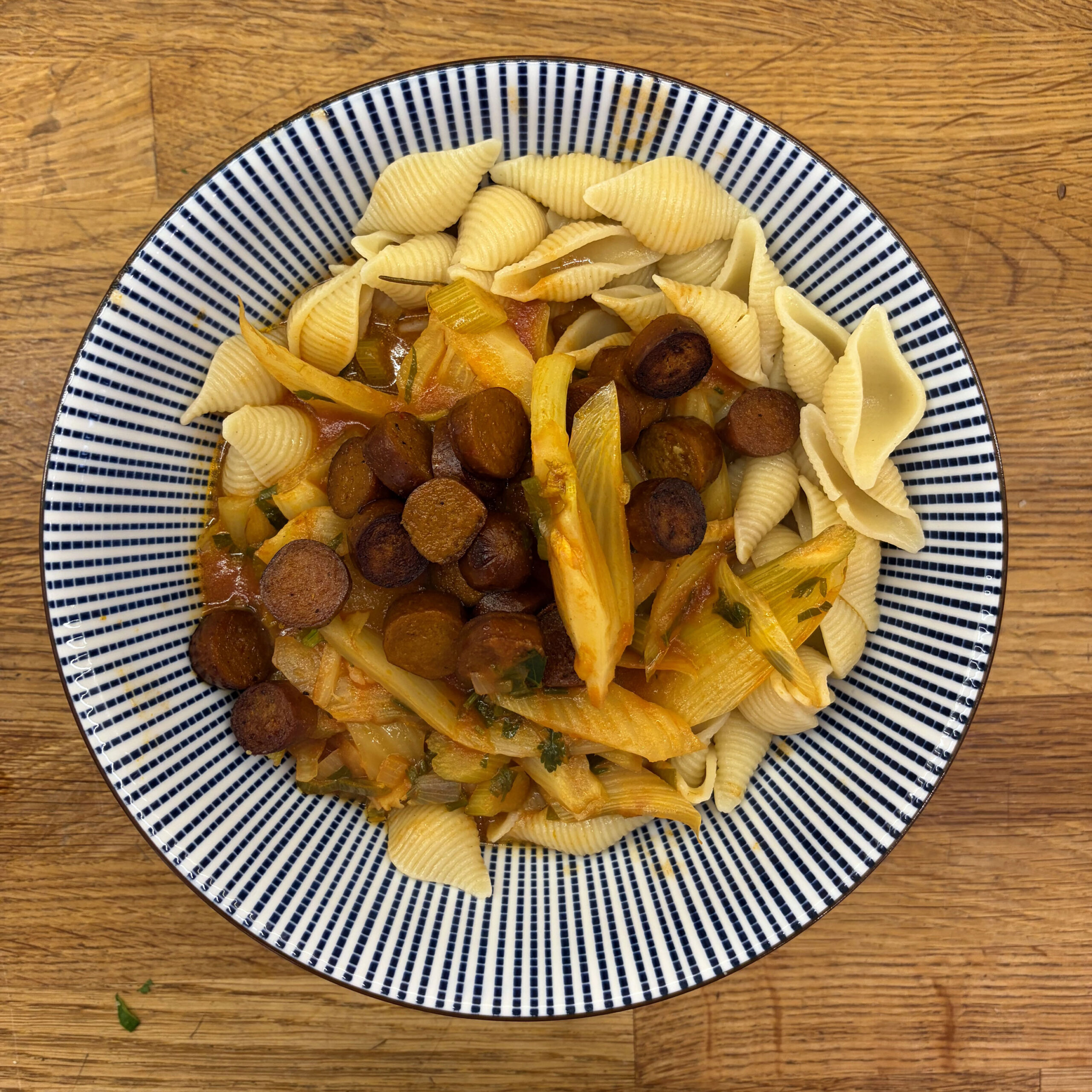 Teller mit Conchiglie-Muschelnudeln, Fenchel-Tomaten-Ragout und gebratenen Scheiben veganer Merguez auf blau-weiß gestreiftem Teller.