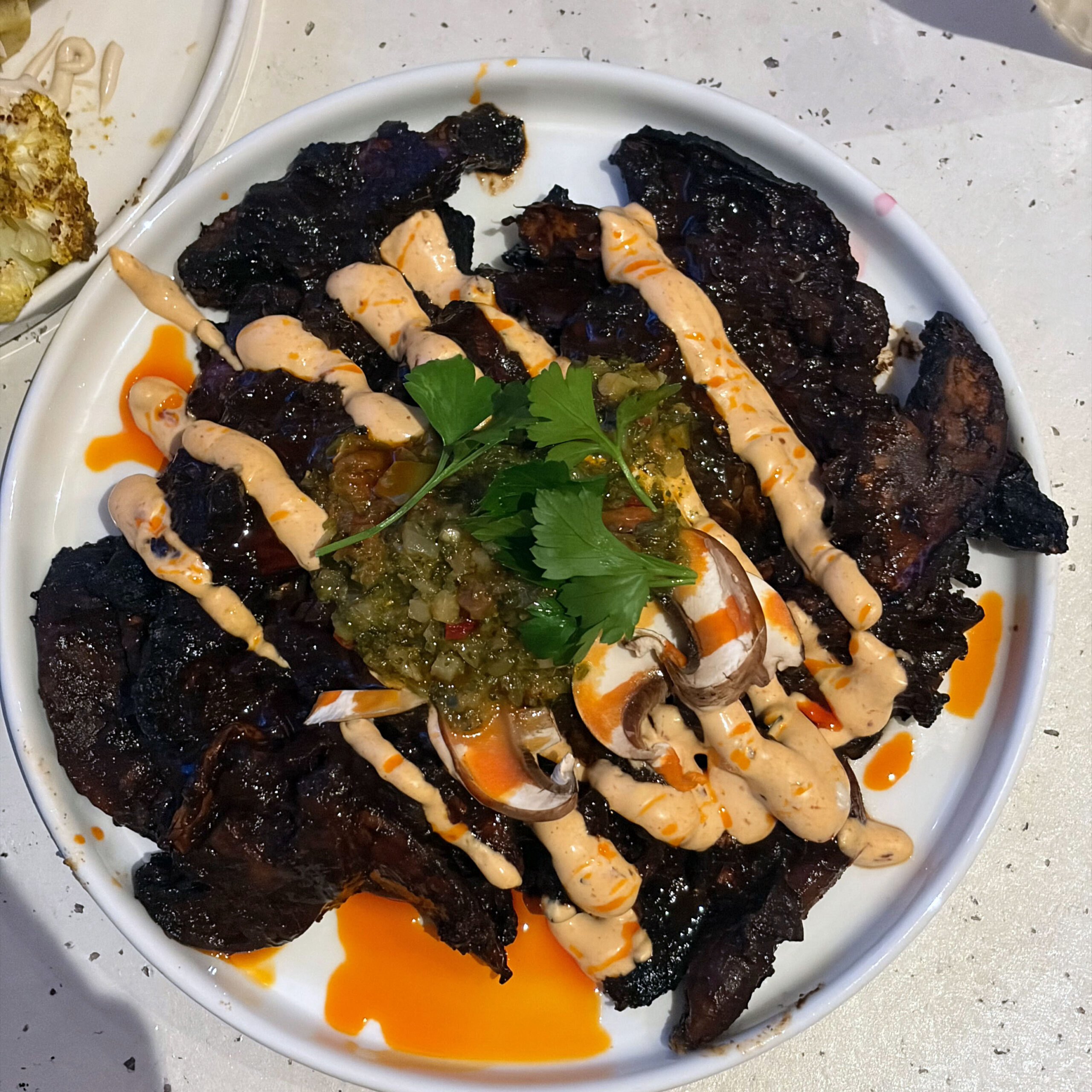BBQ-gegrillte Pilze auf weißem Teller, garniert mit grüner Salsa, frischem Koriander, cremiger Chipotle-Sauce und orangefarbenem Chiliöl.