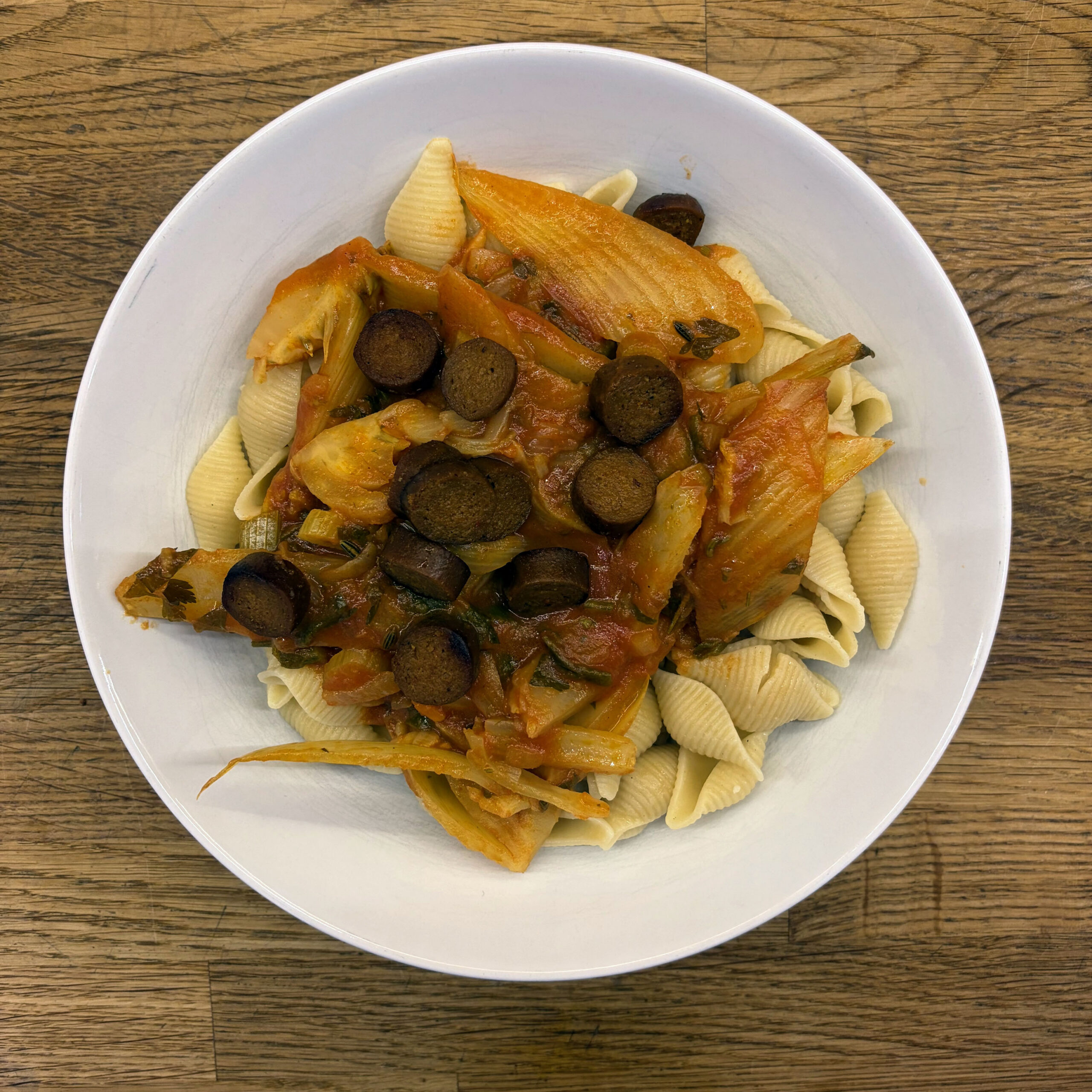 Weißer tiefer Teller mit Conchiglie-Nudeln, belegt mit Fenchel-Ragout in Tomatensauce und gebratenen Merguez-Scheiben, serviert auf einem Holztisch.