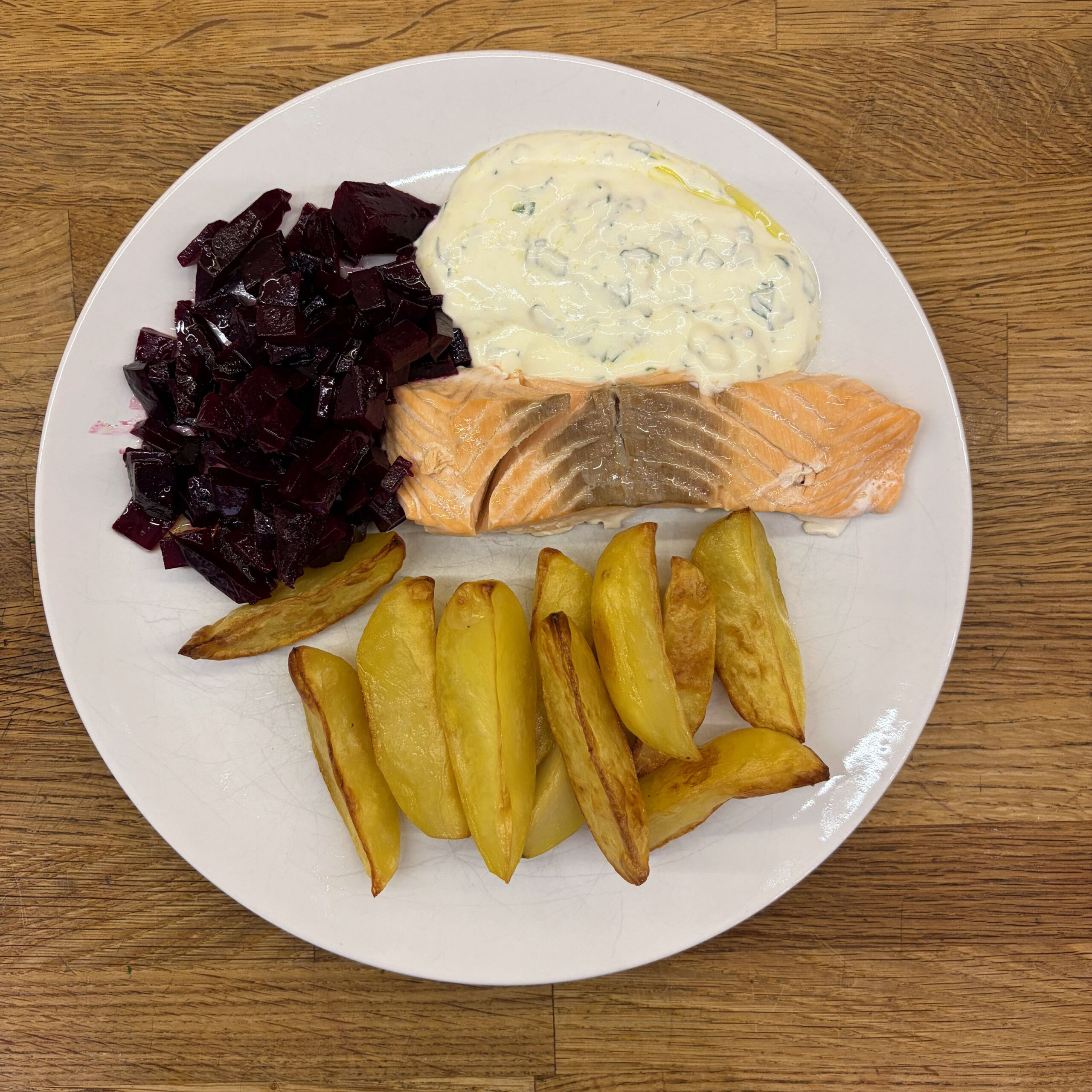 Weißer Teller mit einem gedünsteten Lachsfilet, belegt mit Joghurt-Petersilien-Dipp, daneben gewürfelter Rote-Bete-Salat und knusprige Kartoffelecken aus dem Ofen.