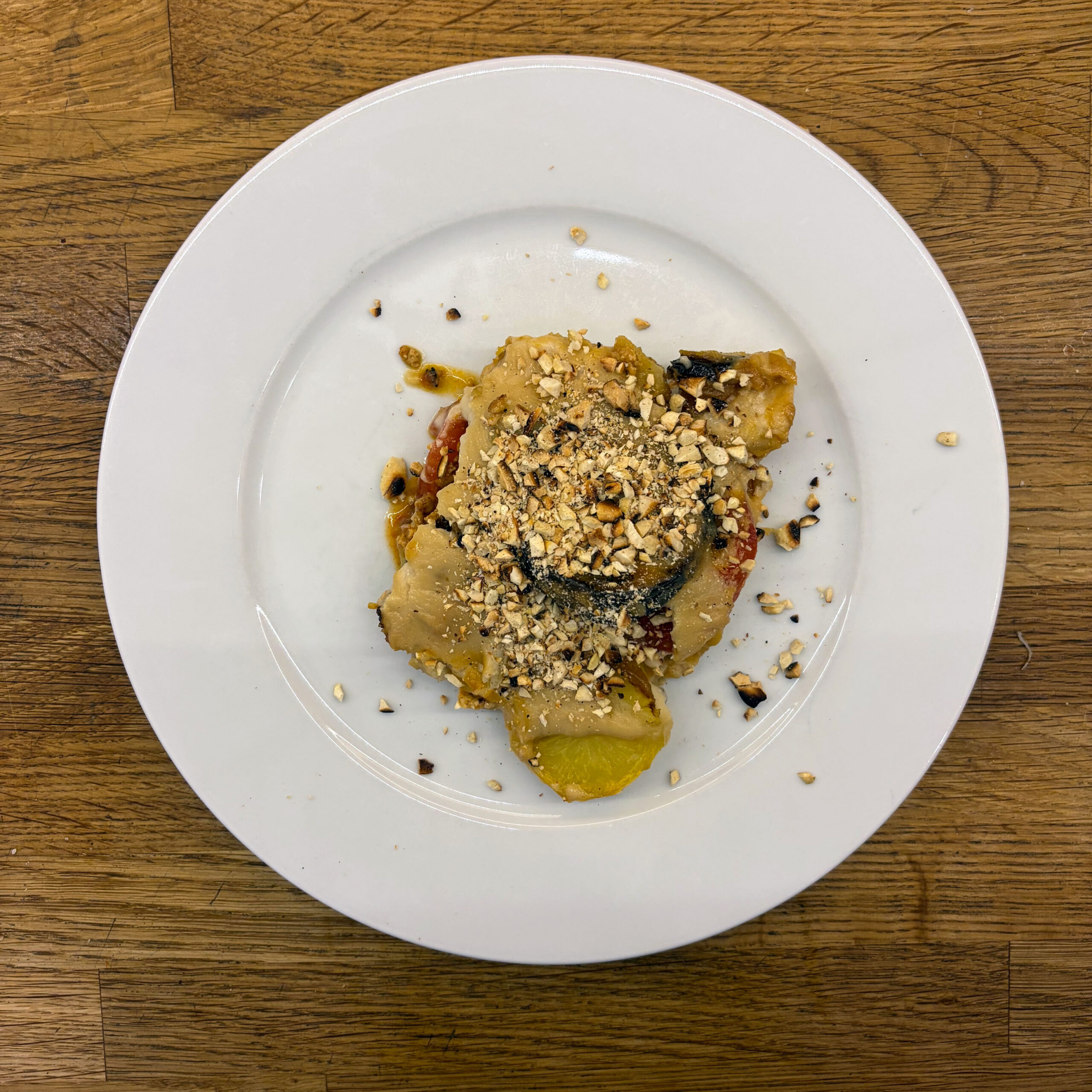 Veganes Moussaka auf einem weißen Teller: Schichten aus Kartoffelscheiben, gebratenen Auberginenscheiben, Tofu-Tomatensauce und veganer Béchamel, garniert mit gehackten gerösteten Mandeln und schwarzem Pfeffer.