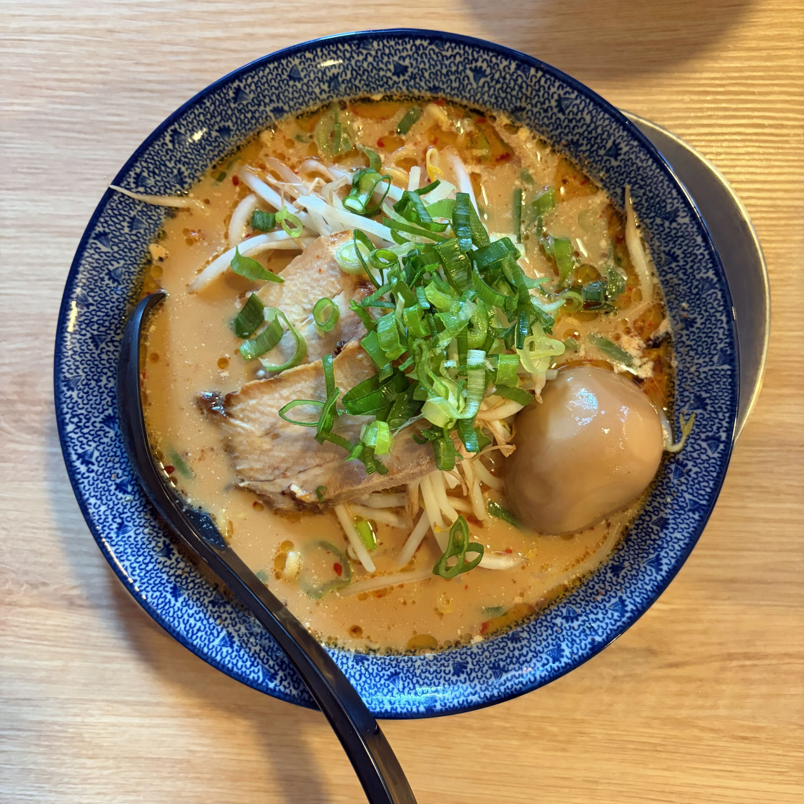 Eine Schüssel Kara-Shoyu-Ramen mit würzig-scharfer Brühe, Chashu-Schweinefleisch, einem Sojasauce-Ei, Sojasprossen, Frühlingszwiebeln und Chili-Flocken in einer blau-weißen Schüssel.