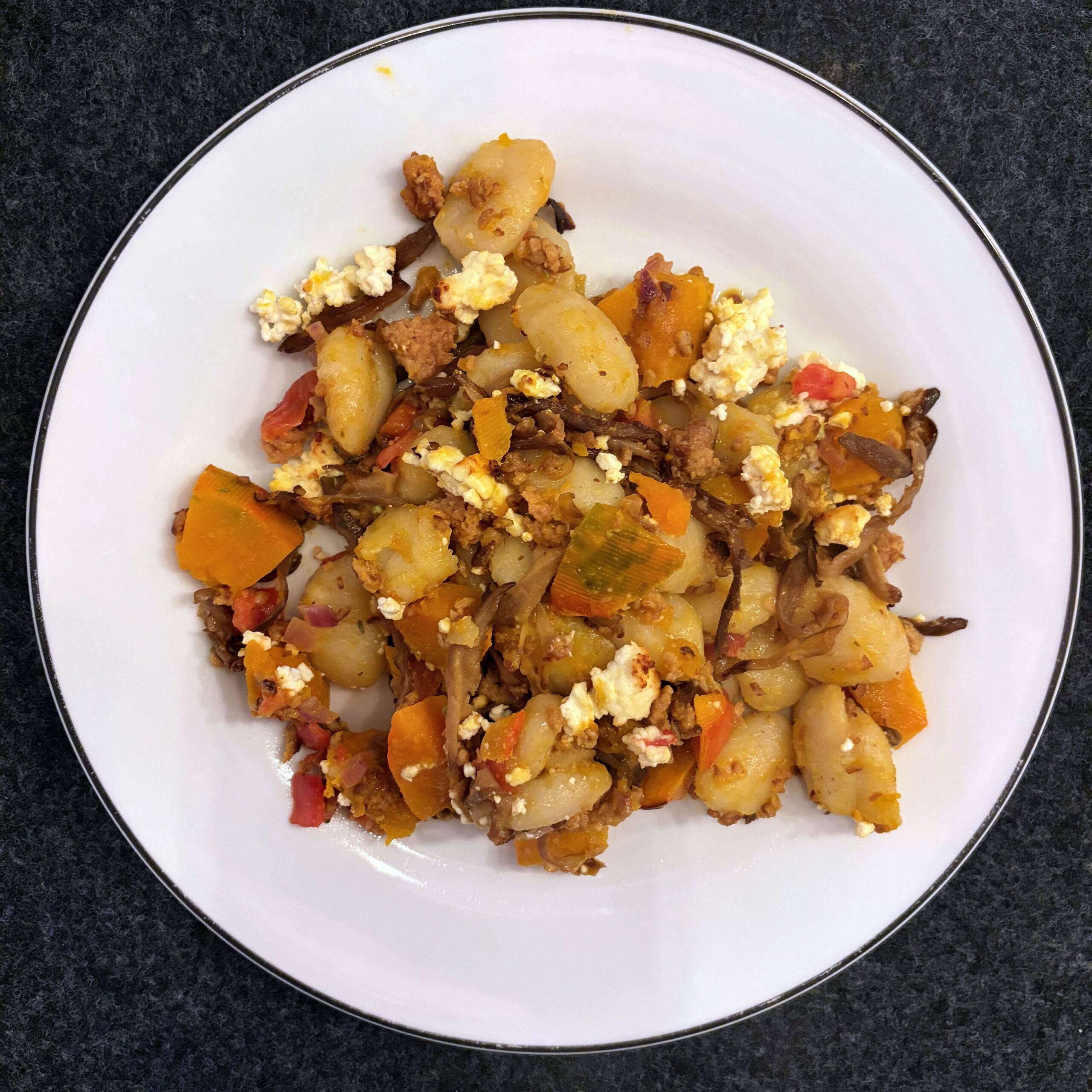 Ein Teller mit gebackenen Gnocchi, Kürbissstücken, Austernpilzen und Veggiehack in Sahnesoße, garniert mit zerkrümeltem Feta-Käse und roten Paprikawürfeln.