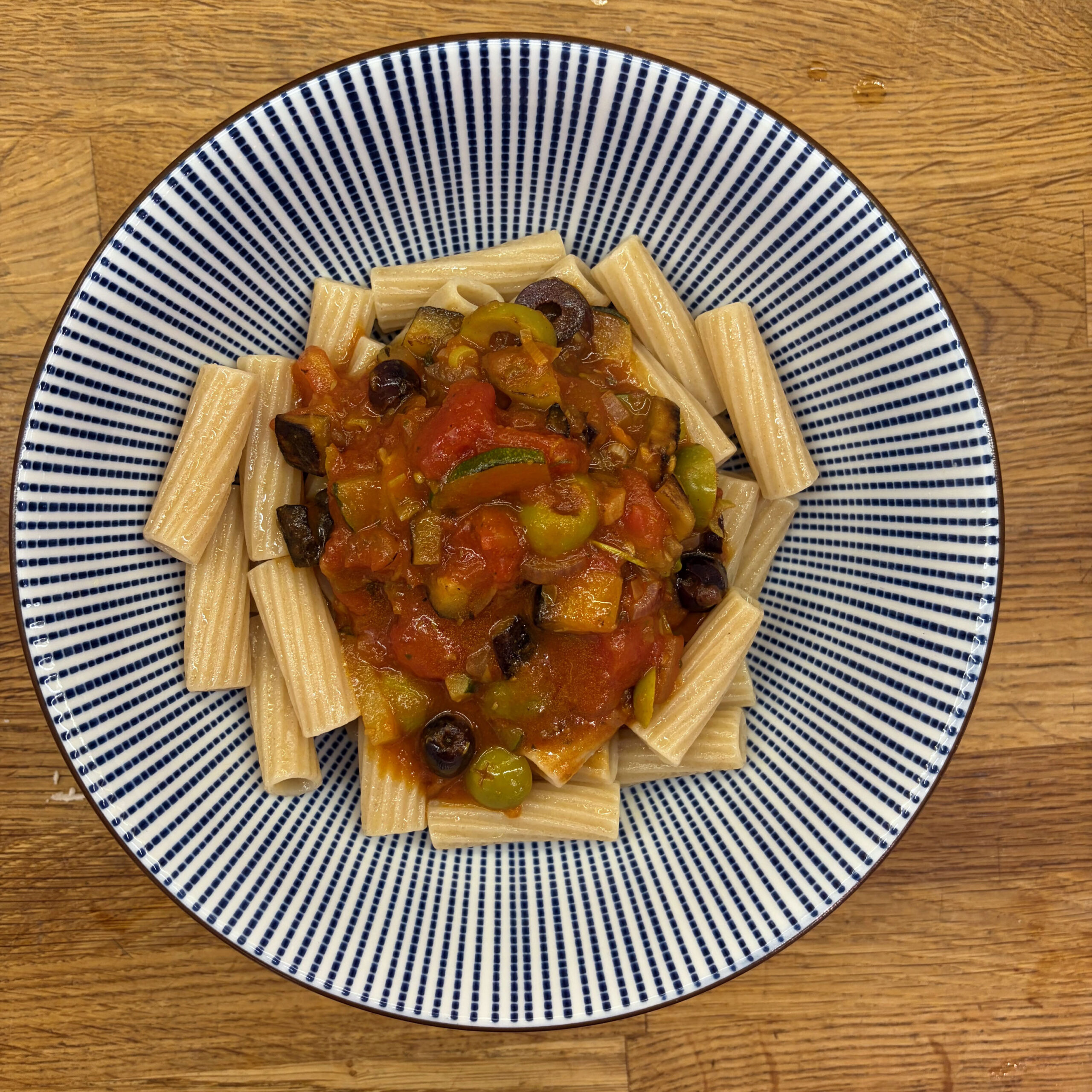 Rigatoni auf einem blau-weiß gestreiften Teller, angerichtet mit einer Tomatensoße aus Aubergine, Zucchini, Oliven und Kapern.